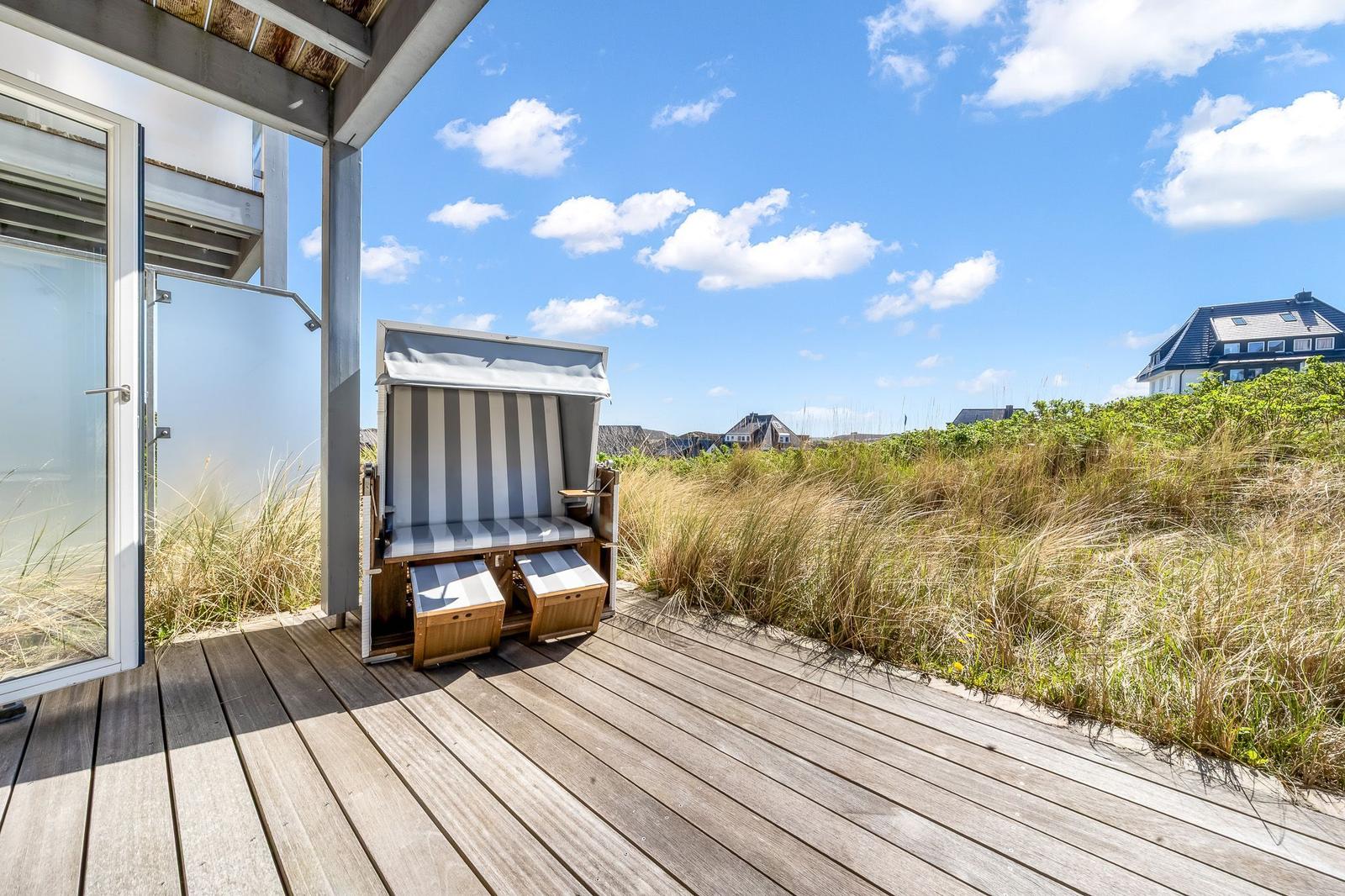 Terrasse mit Strandkorb, Holzdecke und Blick auf Dünen und Häuser.