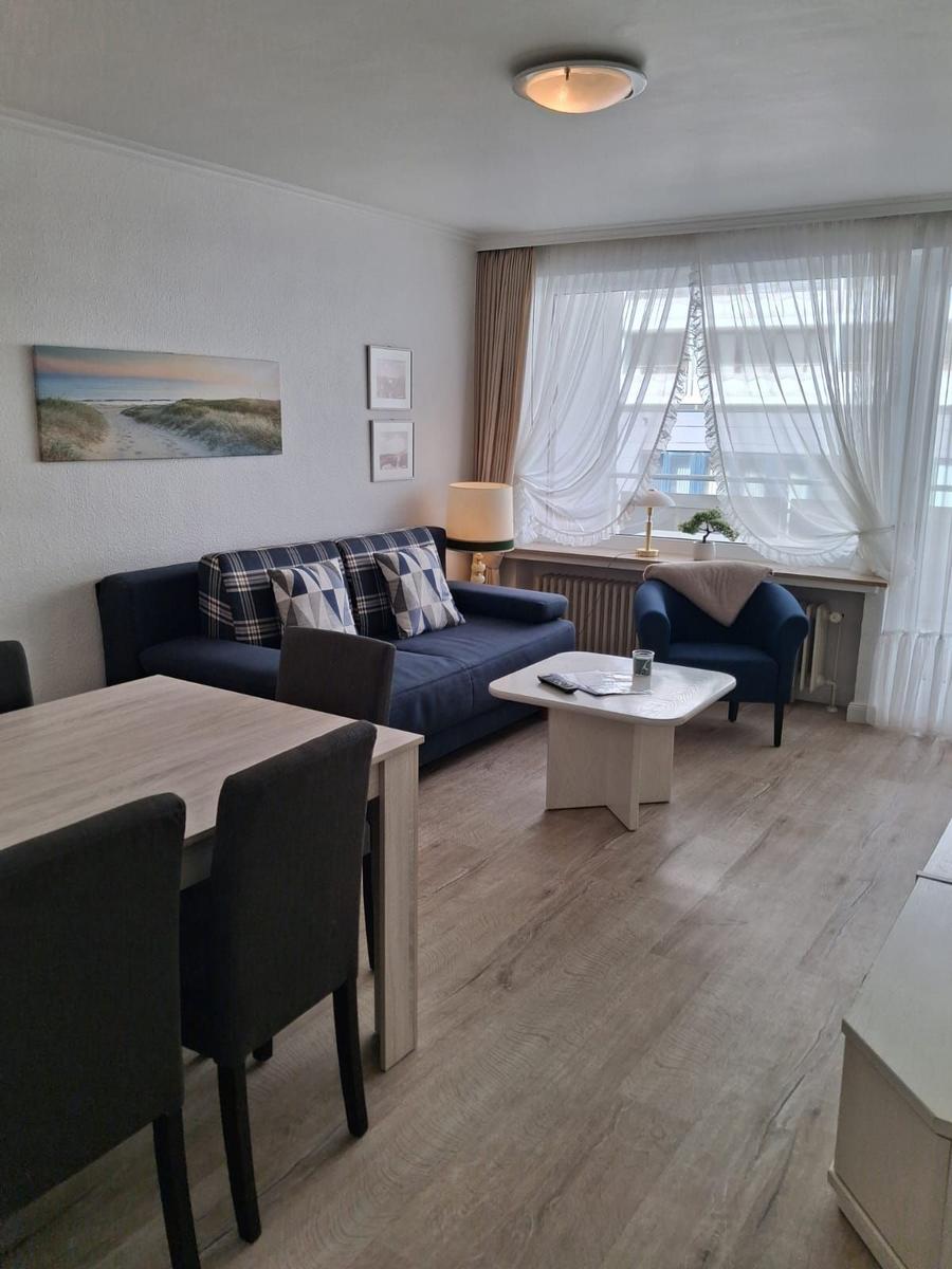 Wohn- und Essbereich mit Sofa, Tisch und Stühlen. Fenster mit Vorhängen und Wandbild.