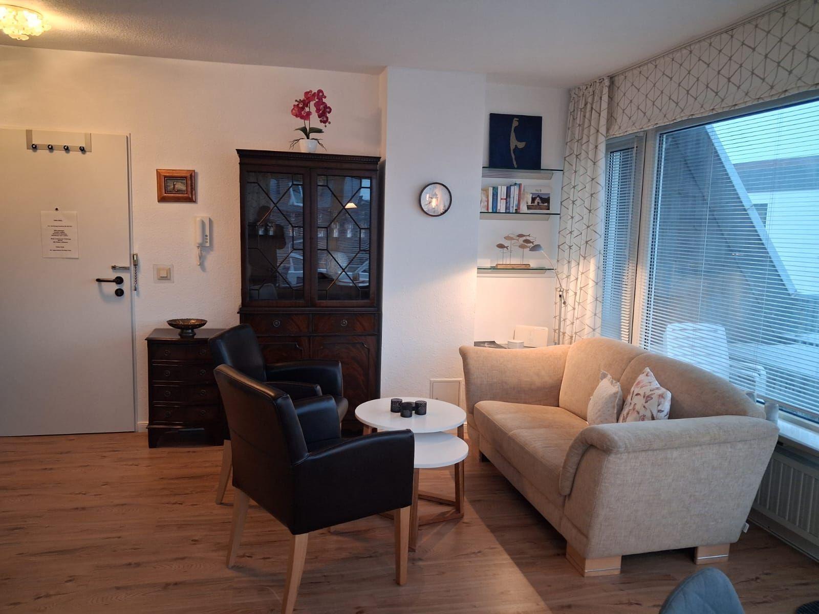 Living room with sofa, two chairs, and glass-front cabinet. Windows with blinds and wooden floor.