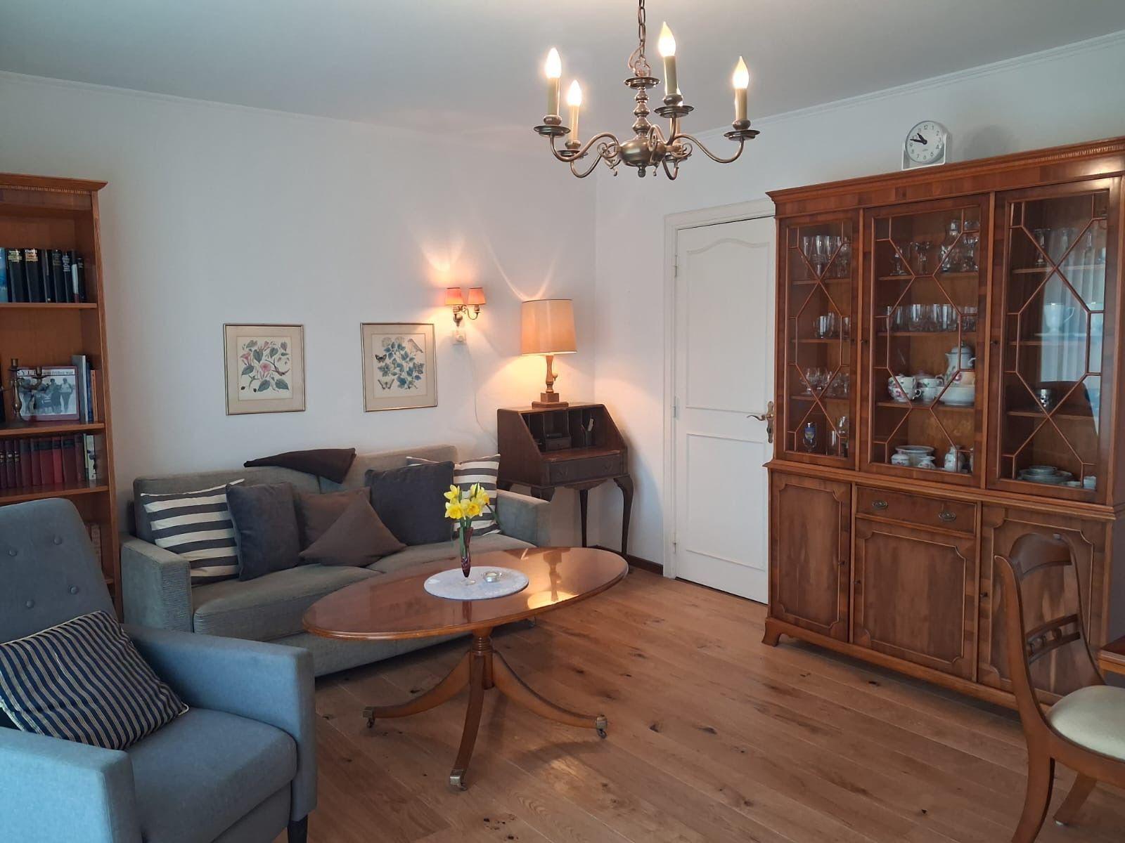 Living room with sofa, armchair, table, and wooden cabinet. Chandelier and bookcase visible.