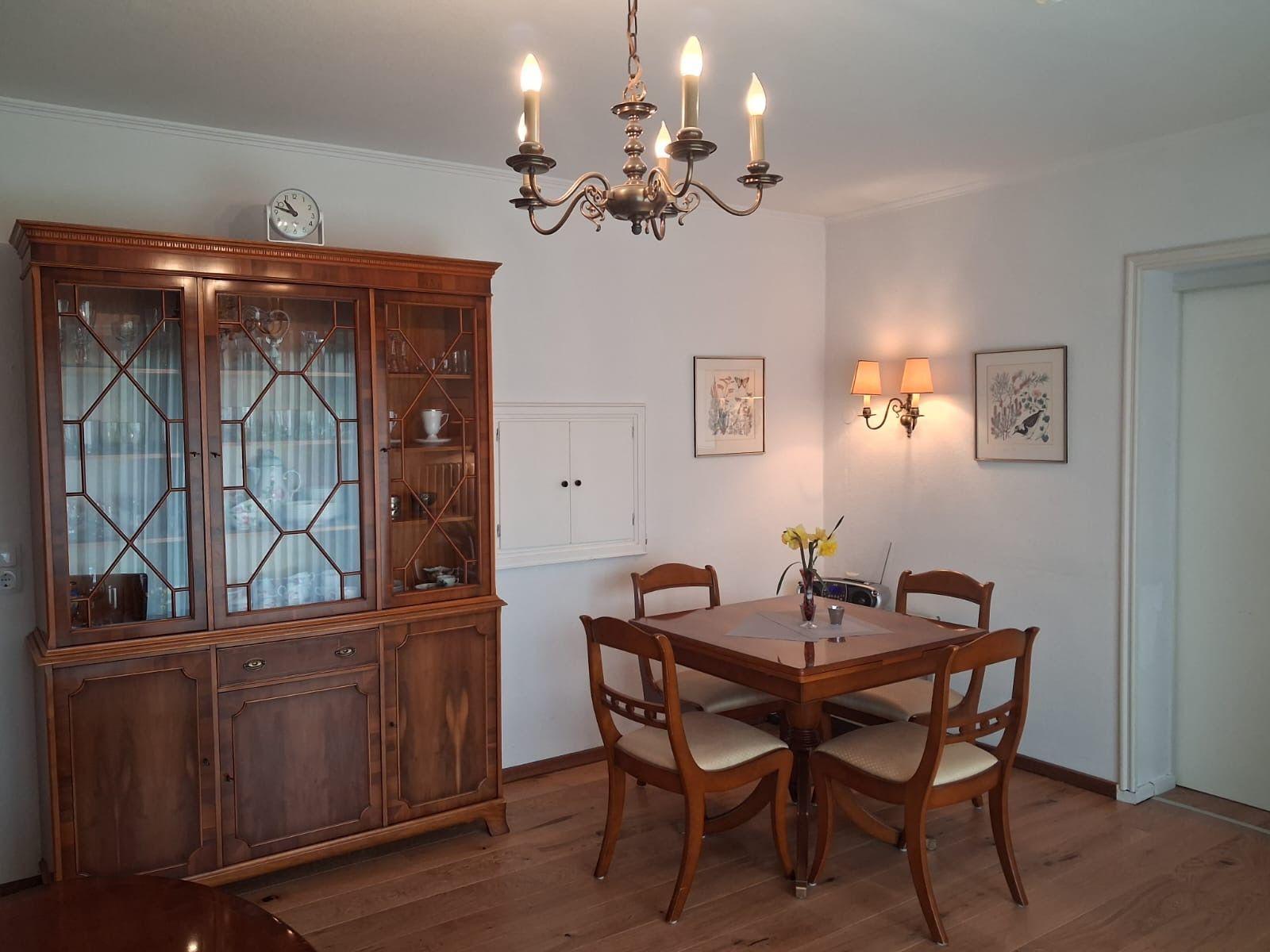 Dining room with wooden table, four chairs, and a glass display cabinet. Ceiling and wall lamps illuminate the space.