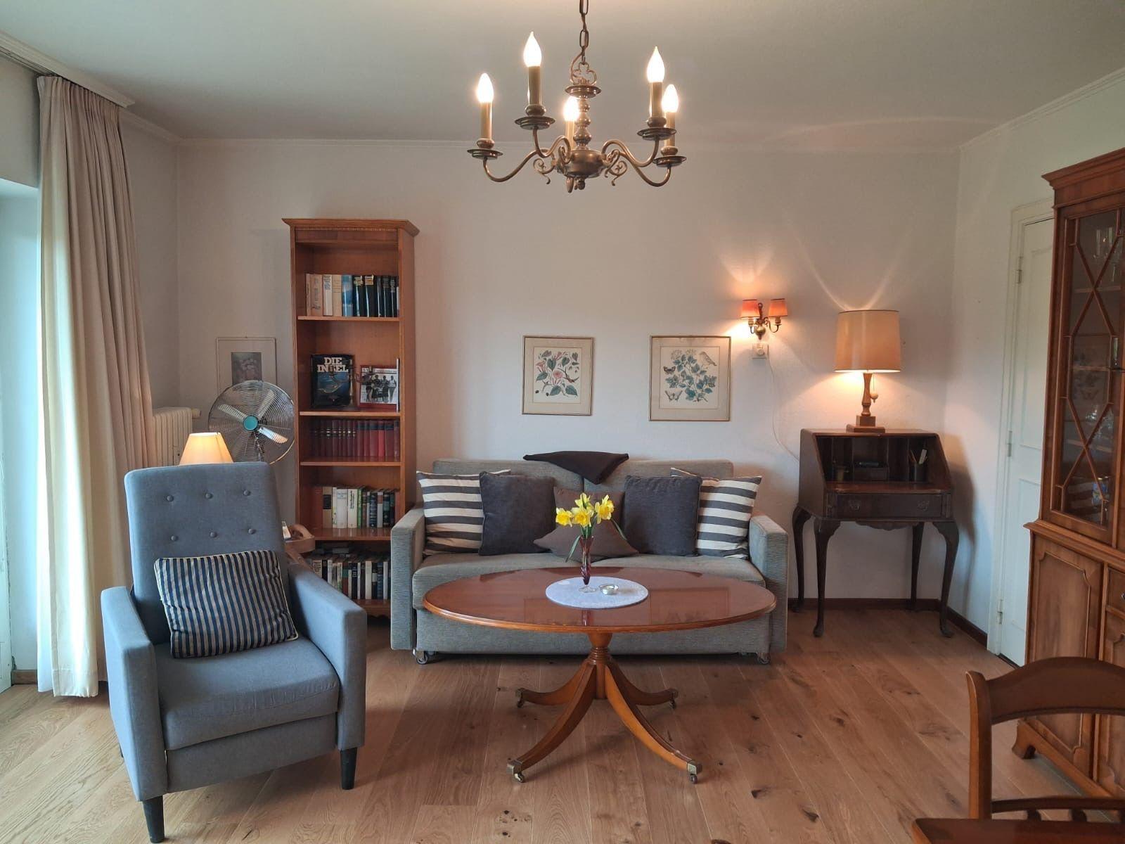 Living room with sofa, armchair, table, and bookcase. Wooden floor and chandelier.
