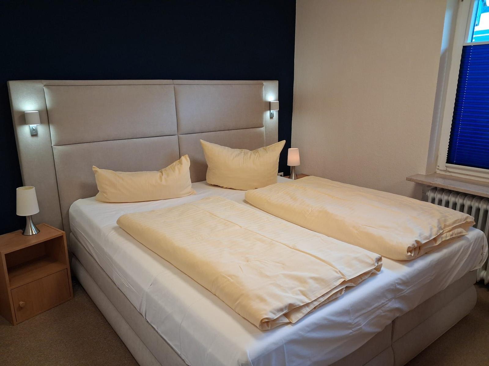 Doppelbett mit Bettwäsche, Nachttisch und Wandlampe. Fenster mit Jalousie.