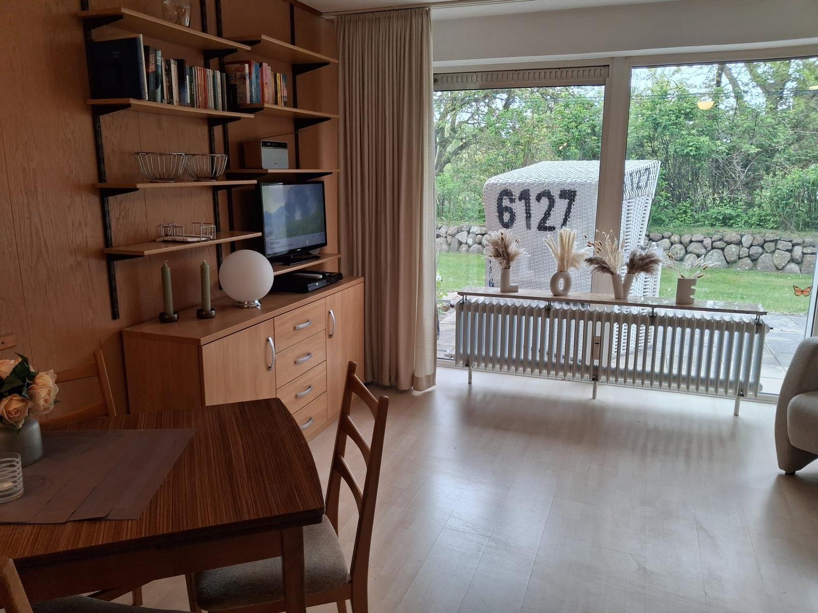 Essbereich mit Tisch und Stühlen, Fernseher und Bücherregal. Blick auf Garten mit Sitzmöbel.