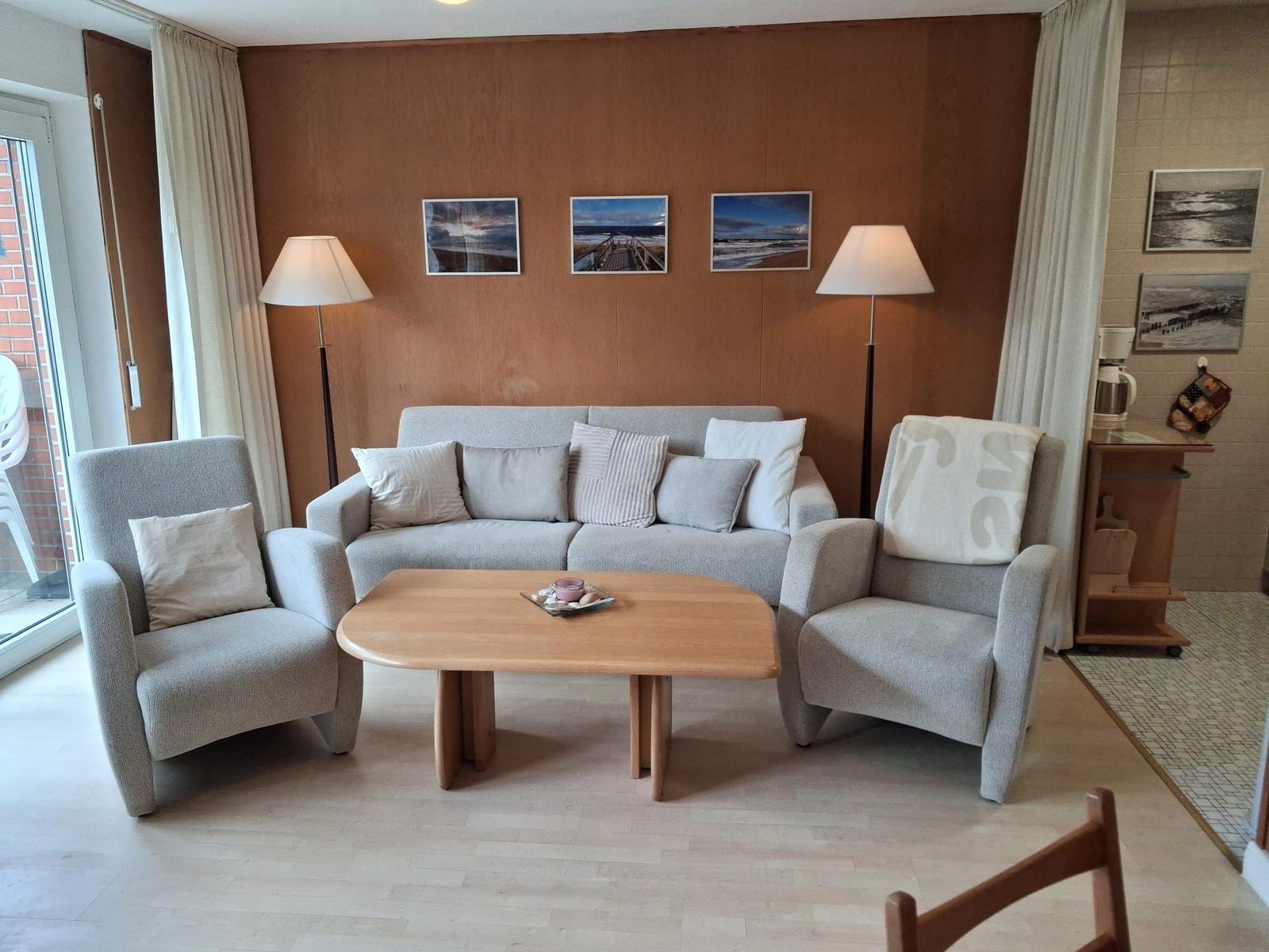 Wohnzimmer mit Sofa, zwei Sesseln und Holztisch. An der Wand hängen drei Bilder.