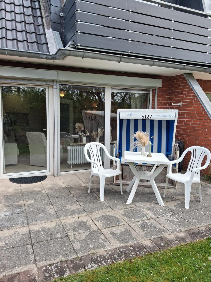 Terrasse mit weißem Tisch und zwei Stühlen, Strandkorb und Glasfront zur Wohnung.