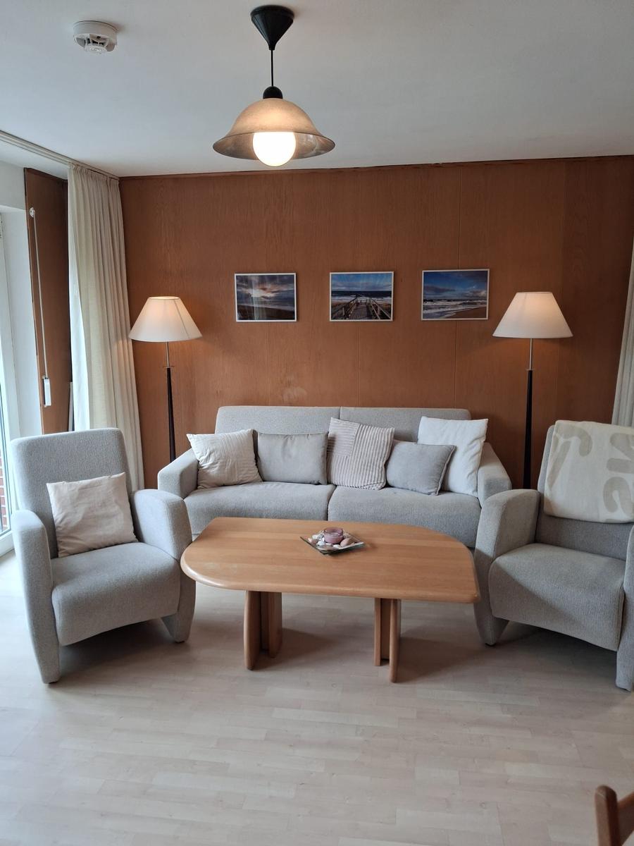Wohnzimmer mit Sofa, zwei Sesseln und Holztisch. Holzwand mit drei Bildern.