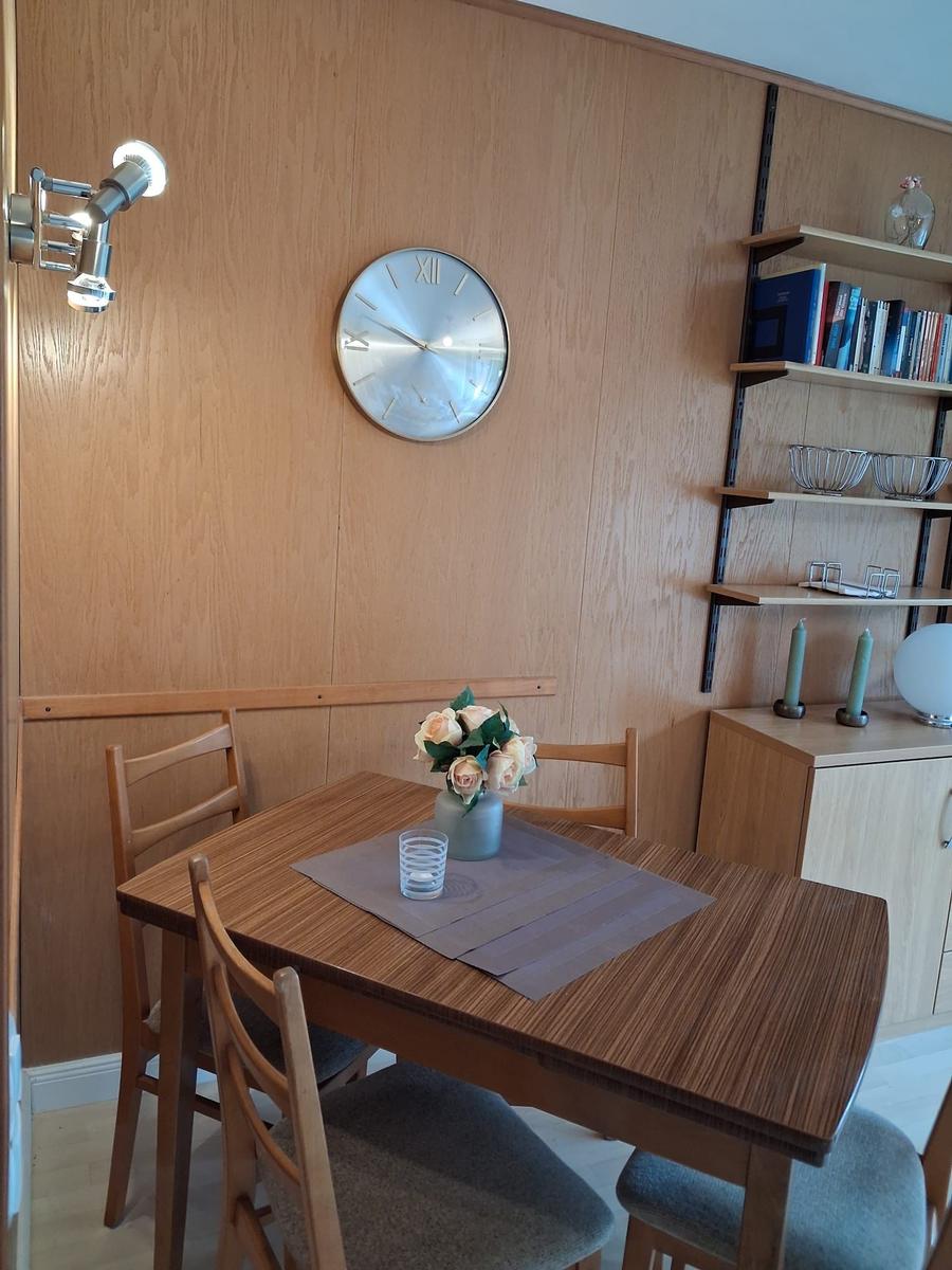 Essbereich mit Holztisch, Stühlen und Wanduhr. Bücherregal und Blumen auf Tisch.