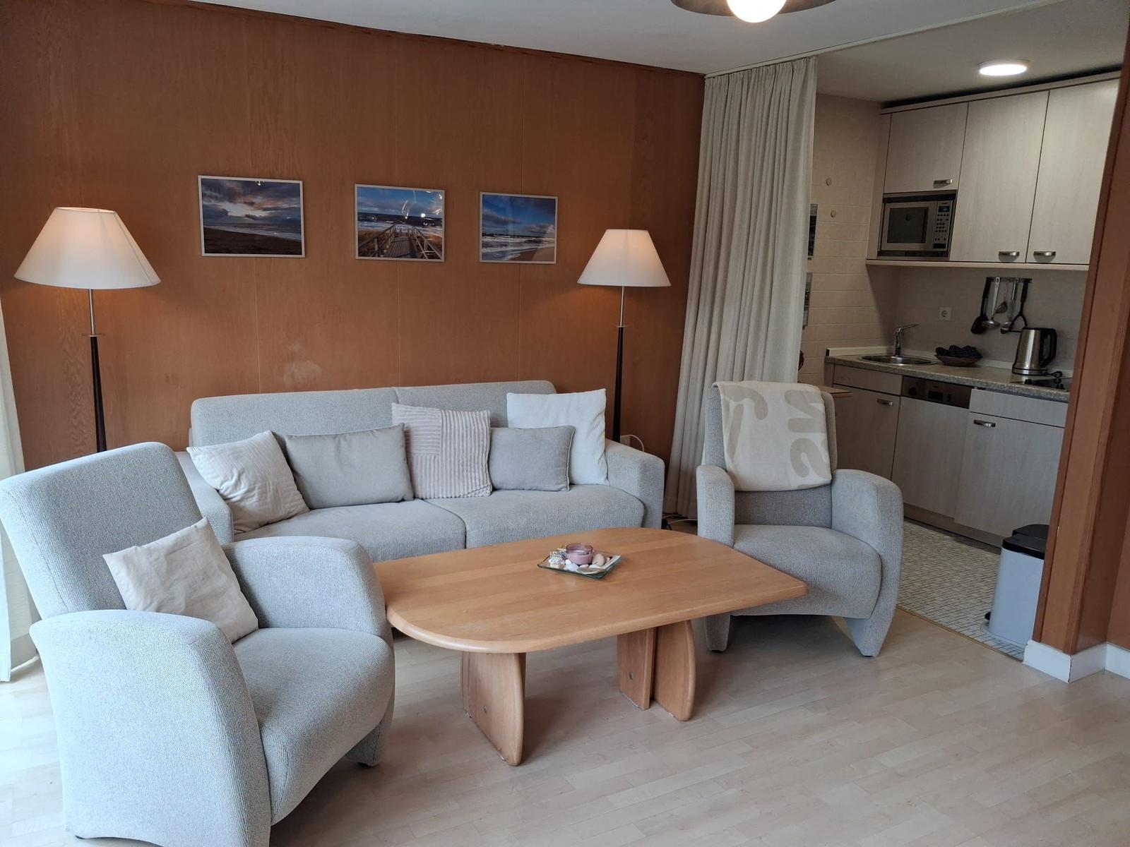 Wohnbereich mit Sofa, zwei Sesseln und Holztisch. Küche im Hintergrund.