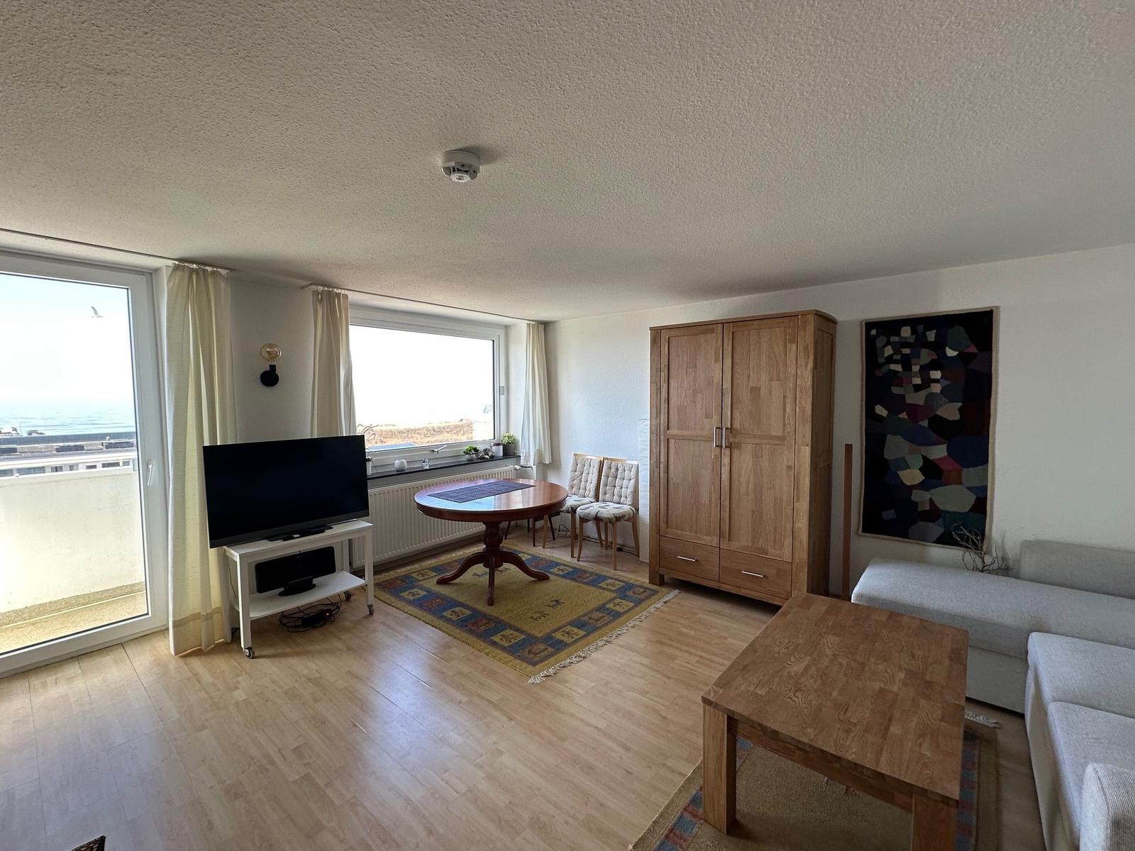 Wohnzimmer mit Sofa, Tisch, Schrank und Fernseher. Fenster und Balkon mit Meerblick.