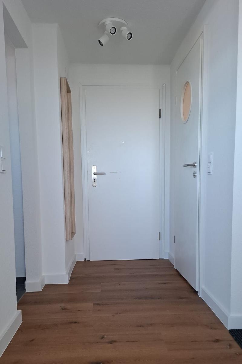 Flur mit Holzboden, weißen Wänden und zwei Türen. Deckenleuchte und runder Spiegel sichtbar.