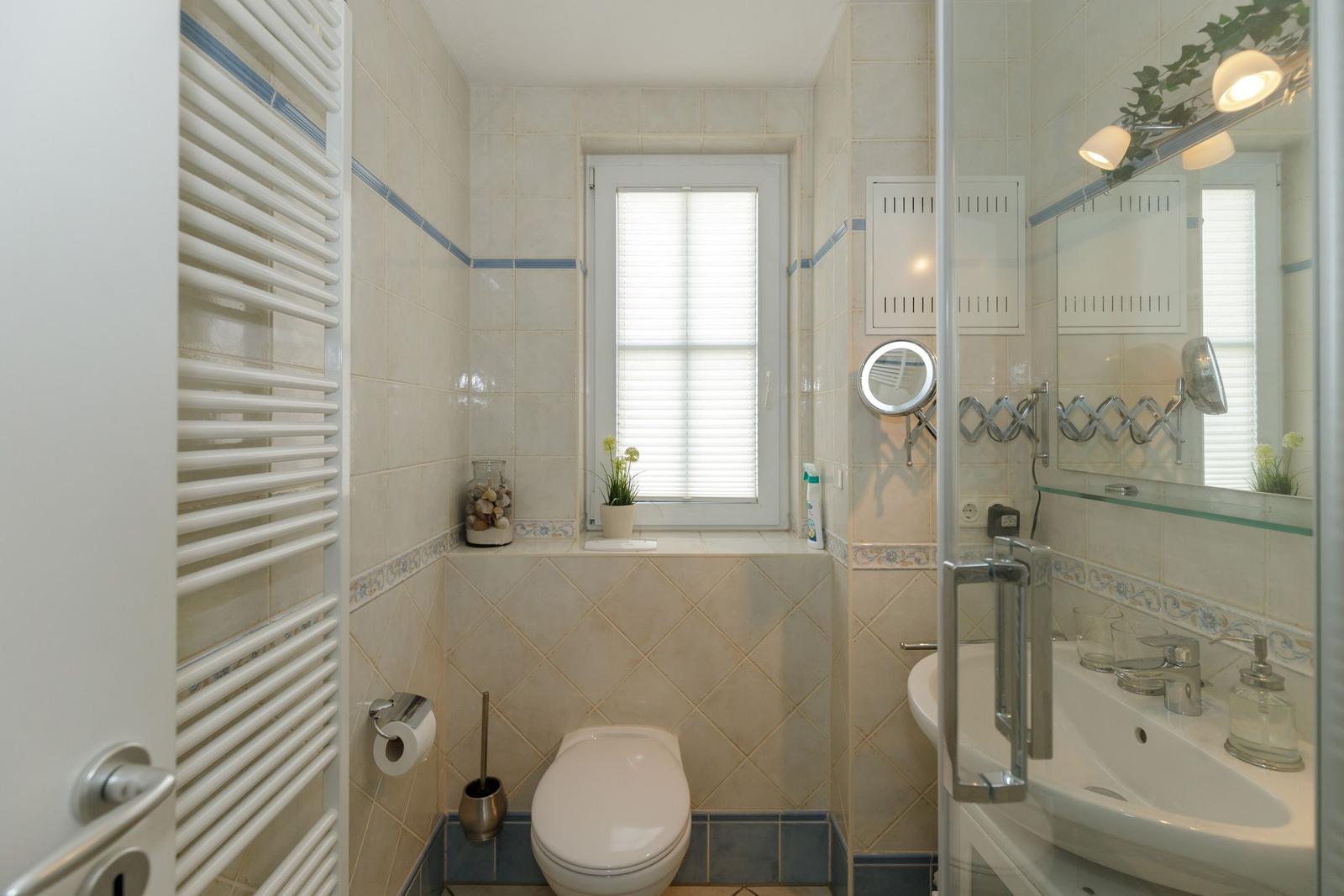 Badezimmer mit Toilette, Waschbecken und Dusche. Fenster mit Jalousie und Pflanze.