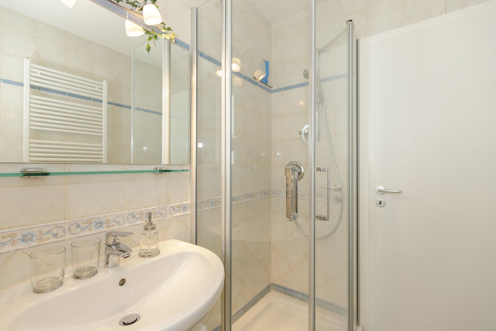 Badezimmer mit Dusche, Waschbecken und Spiegel. Fliesen und Handtuchhalter sichtbar.