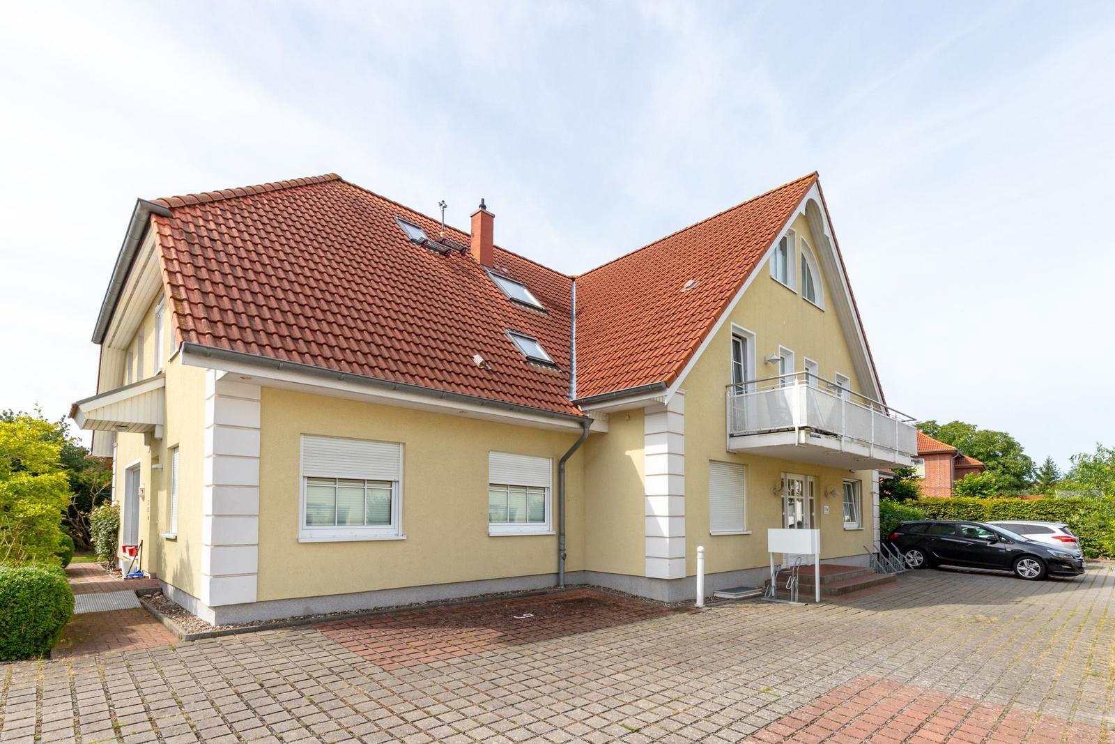 Ein zweistöckiges Haus mit rotem Dach und gelber Fassade. Vor dem Haus befindet sich ein gepflasterter Hof.