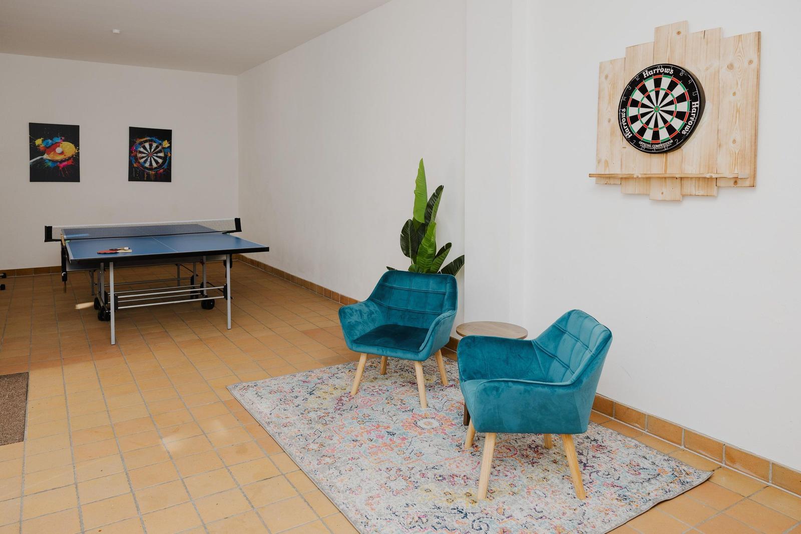 Raum mit Tischtennisplatte, Dartscheibe und zwei blauen Sesseln auf Teppich.