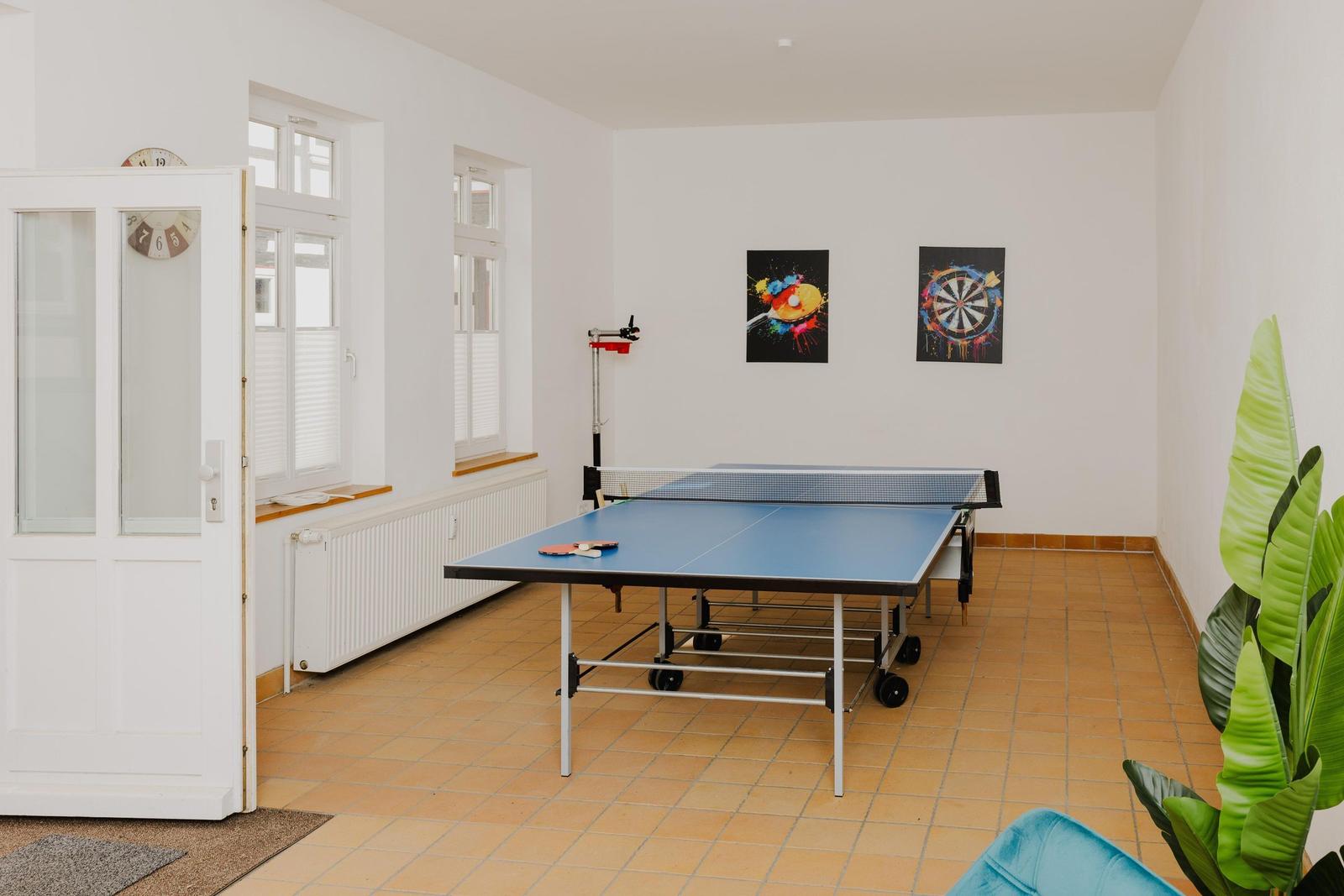 Raum mit Tischtennisplatte, Dartboard und Wandbildern. Tür, Fenster und Pflanze sichtbar.