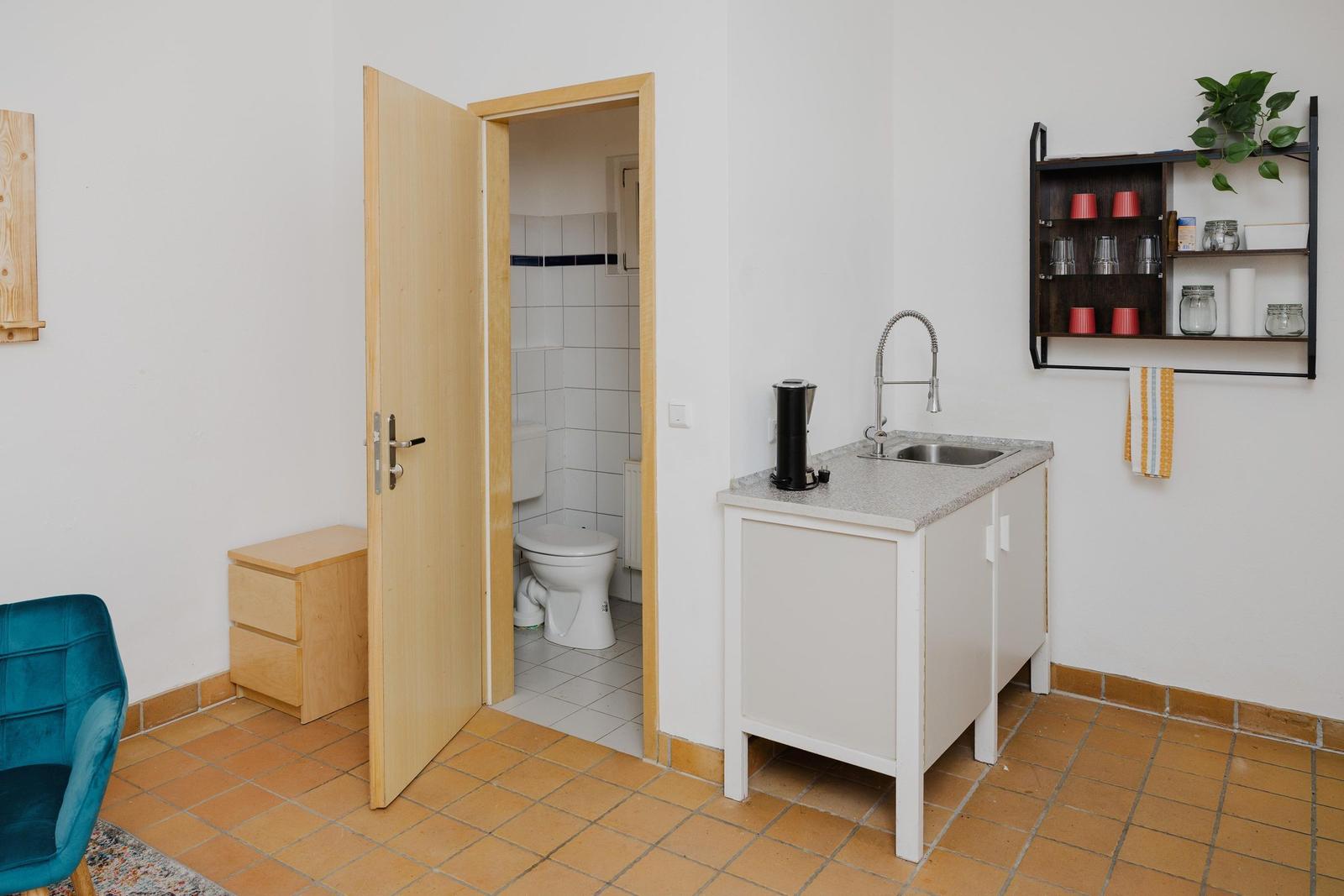 Küche mit Spüle und Kaffeemaschine. Tür führt in die Toilette. Wandregal mit Geschirr und Pflanze.