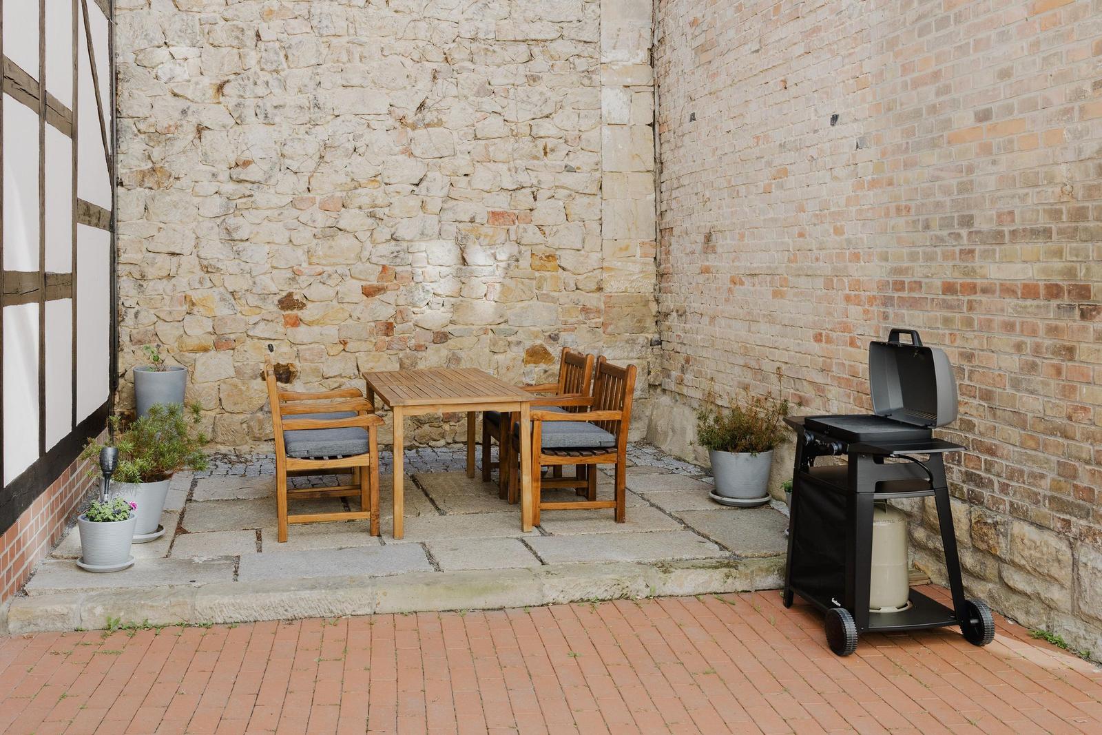 Außenbereich mit Tisch, Stühlen, Grill und Pflanzen an Stein- und Ziegelmauern.