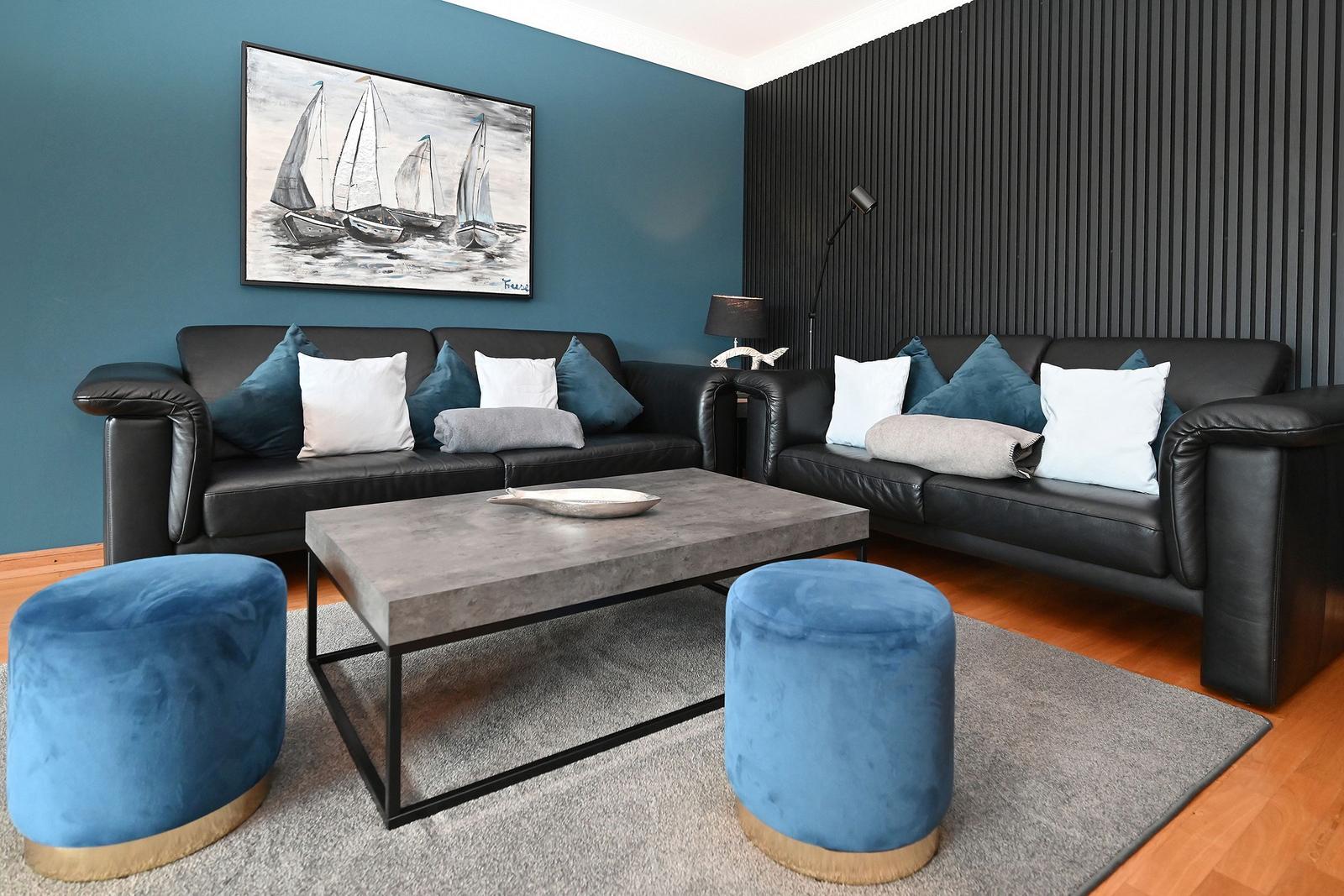 Zwei schwarze Sofas mit Kissen, blau-weiße Sitzkissen, Holzboden, Wandbild mit Segelbooten.
