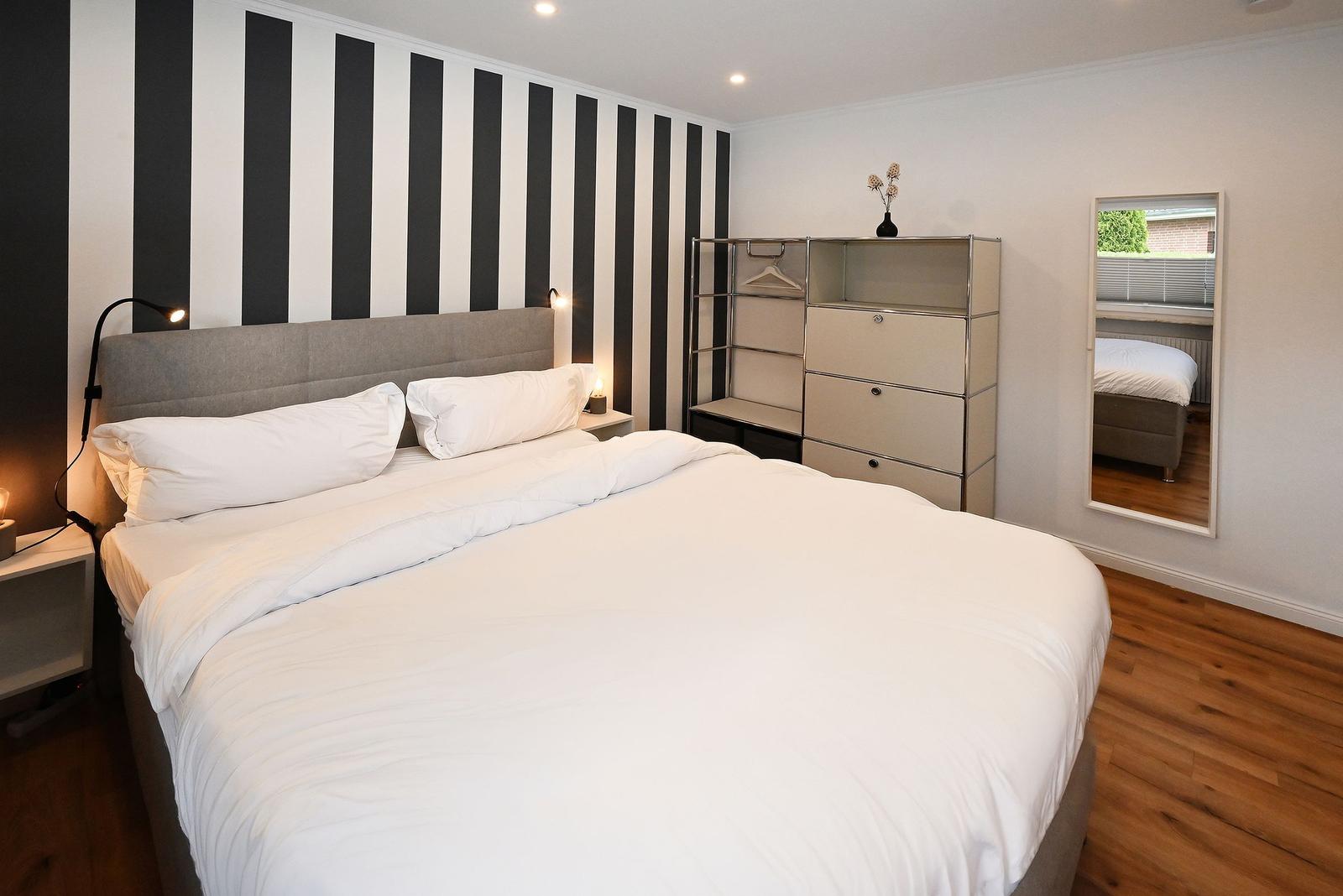 Schlafzimmer mit Bett, Kleiderschrank und Spiegel. Wand mit schwarz-weißen Streifen.