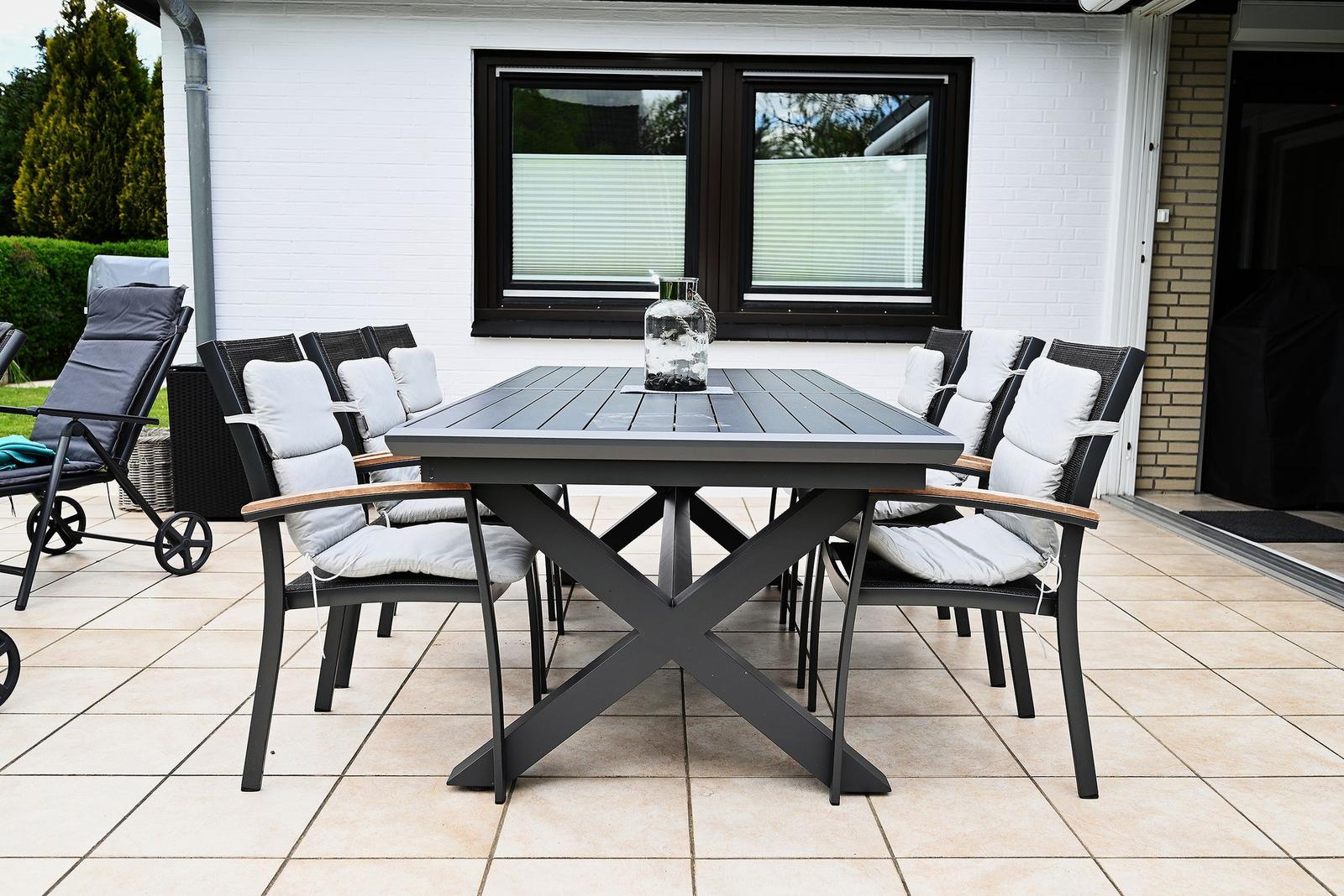 Terrasse mit Esstisch und Stühlen, Glasvase darauf, weißer Putz und Fenster im Hintergrund.