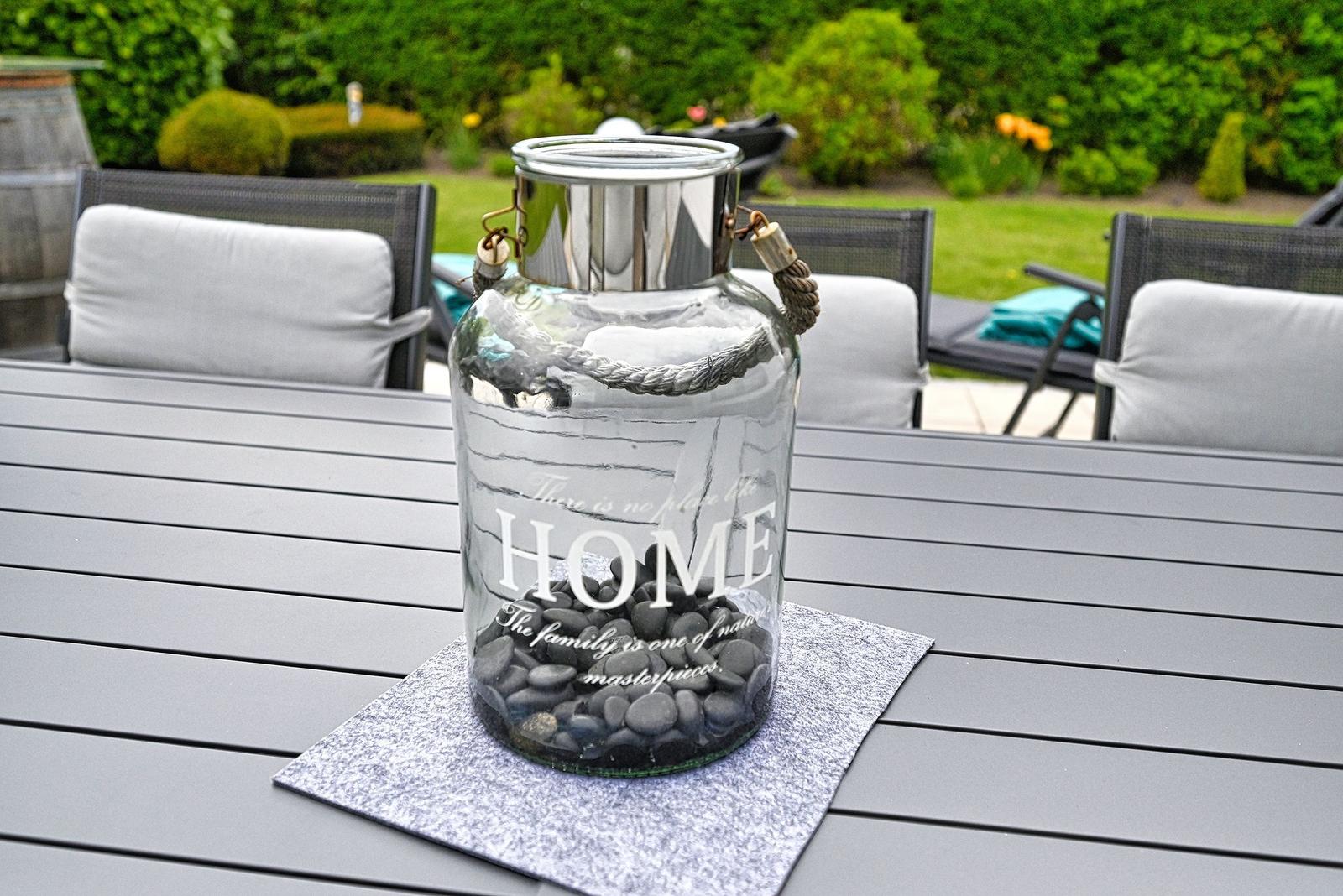 Glasgefäß mit Steinchen und Text steht auf Tisch im Garten. Hintergrund: Gartenmöbel und Grünflächen.