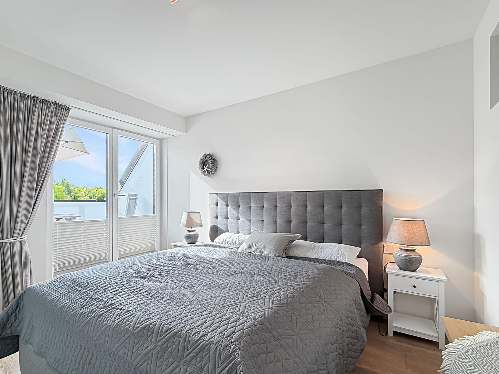 Schlafzimmer mit großem Bett, Fenster zur Terrasse und zwei Nachttischen mit Lampen.