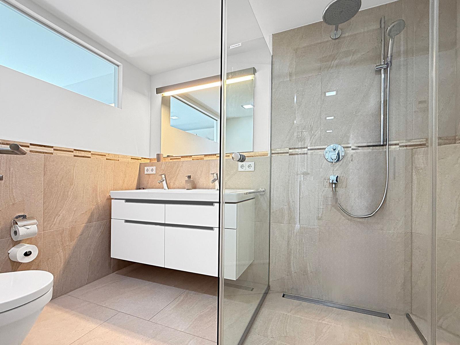 Badezimmer mit Dusche, Waschbecken und Toilette. Große Fenster und Spiegel.