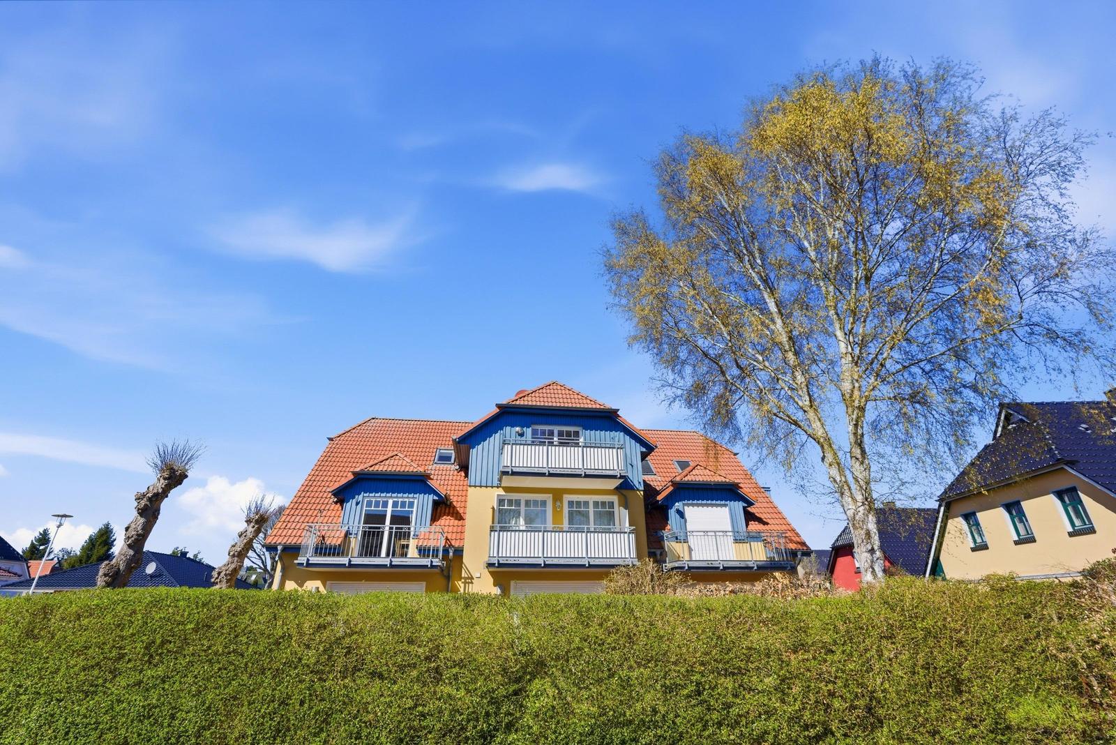 Ein zweistöckiges Haus mit Balkonen und rotem Dach. Grünhecke und Baum im Vordergrund.