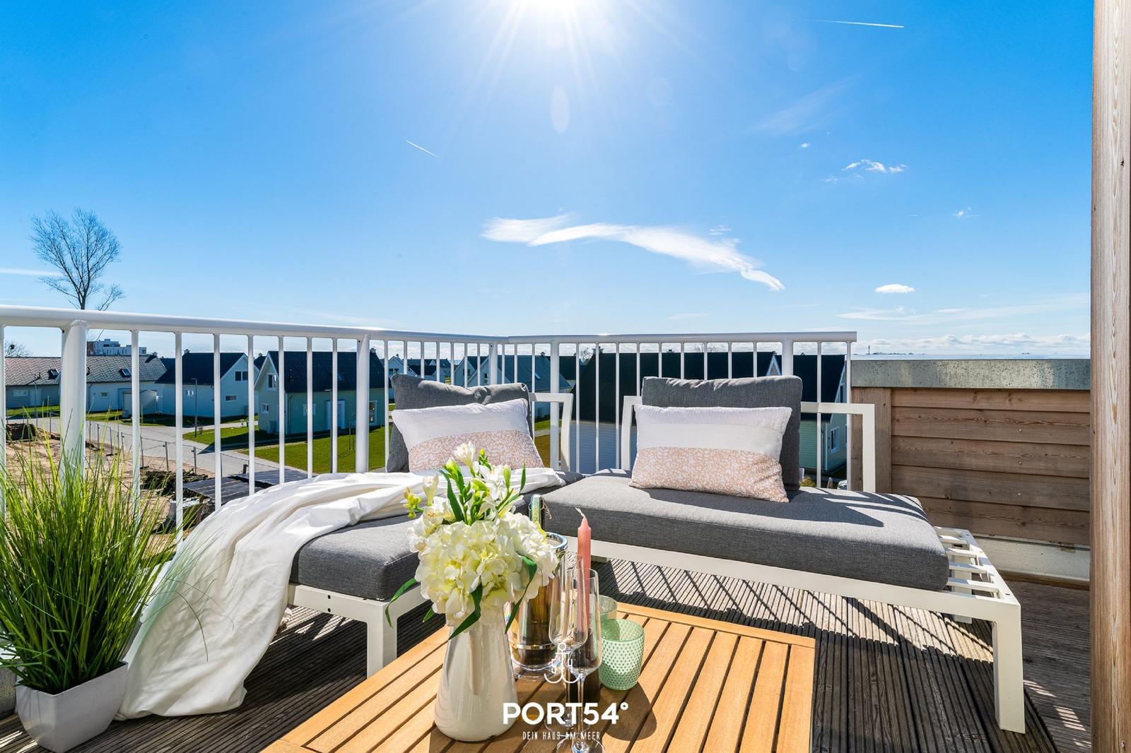 Terrasse mit Liegen, Tisch und Blumen. Blick auf Häuser und Himmel.