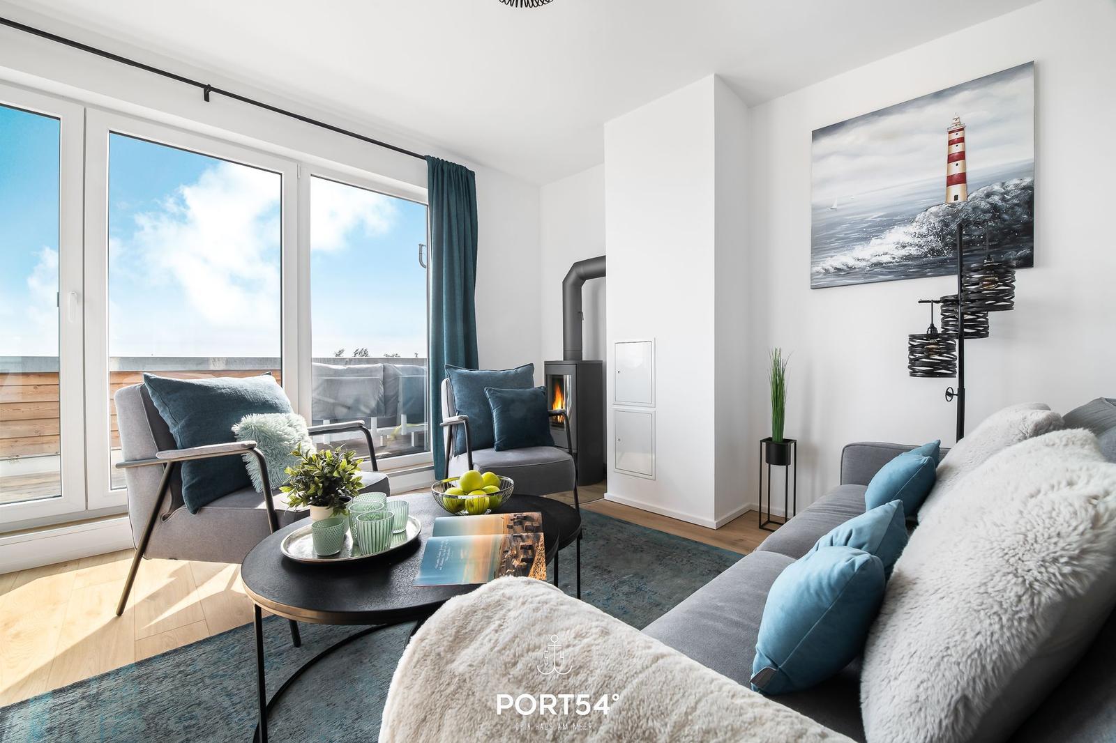 Wohnzimmer mit Sofa, Sessel und Kamin. Große Fenster und Wandbild mit Leuchtturm.