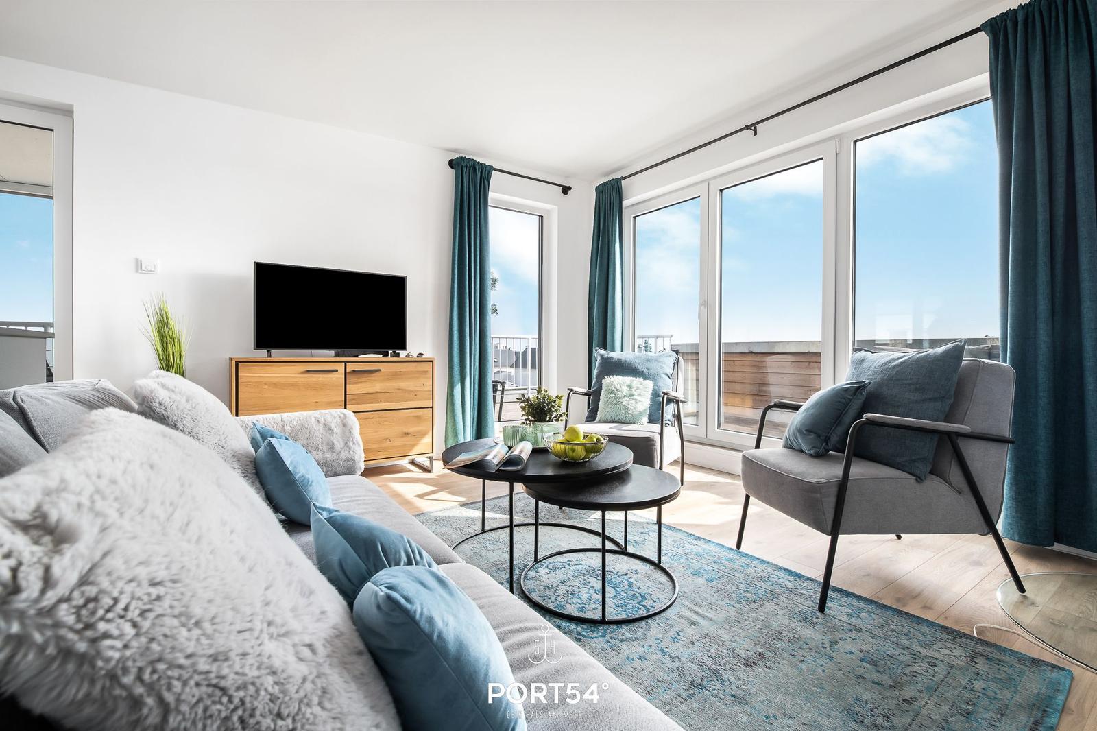 Wohnzimmer mit Sofa, Sesseln und TV. Große Fenster und Balkon.
