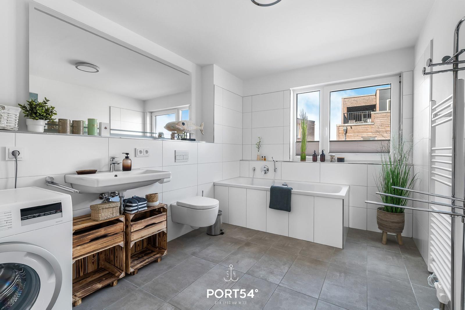 Badezimmer mit Badewanne, Waschmaschine und Holzregal. Fenster mit Blick auf Gebäude.