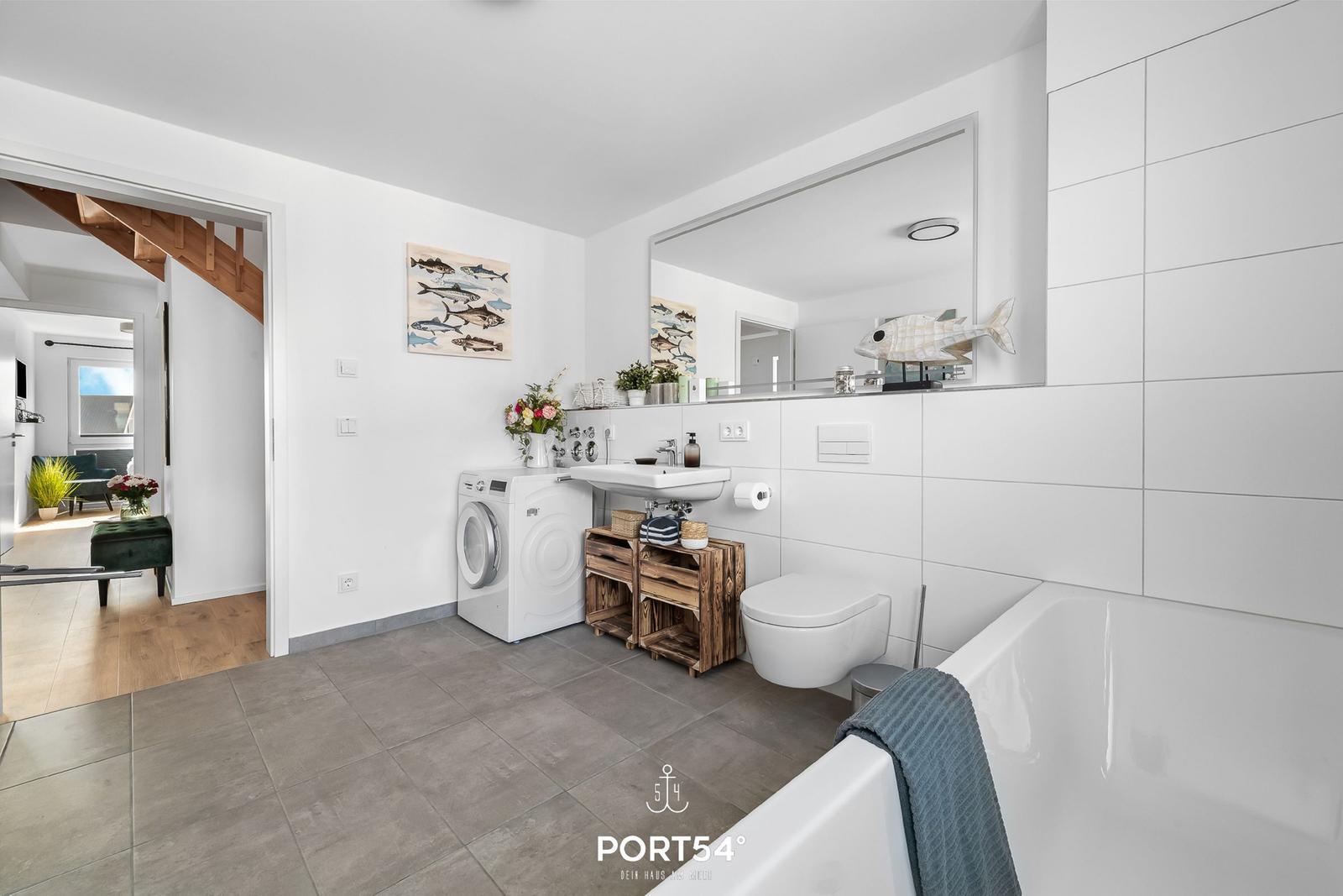 Badezimmer mit Badewanne, Waschmaschine und Spiegel. Holzregal und Wandbild mit Fischen.