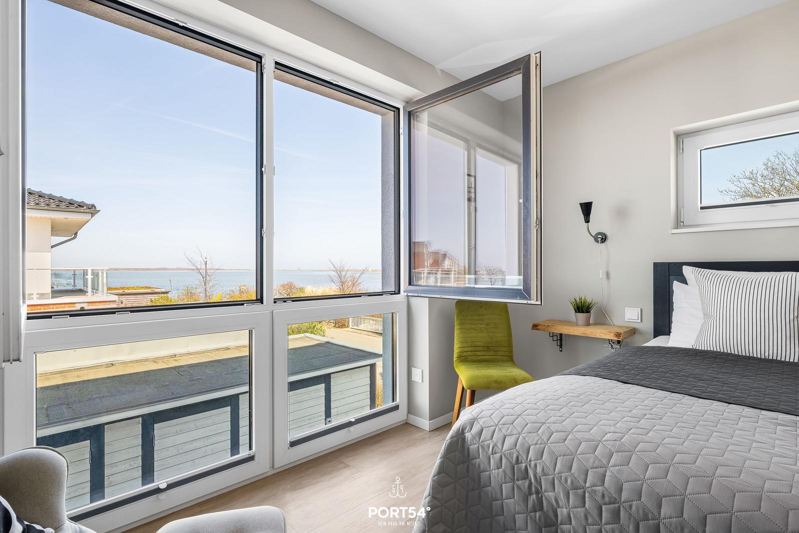 Schlafzimmer mit Blick auf Wasser, großes Fenster, grüne Sitzgelegenheit, Bett mit Kissen.