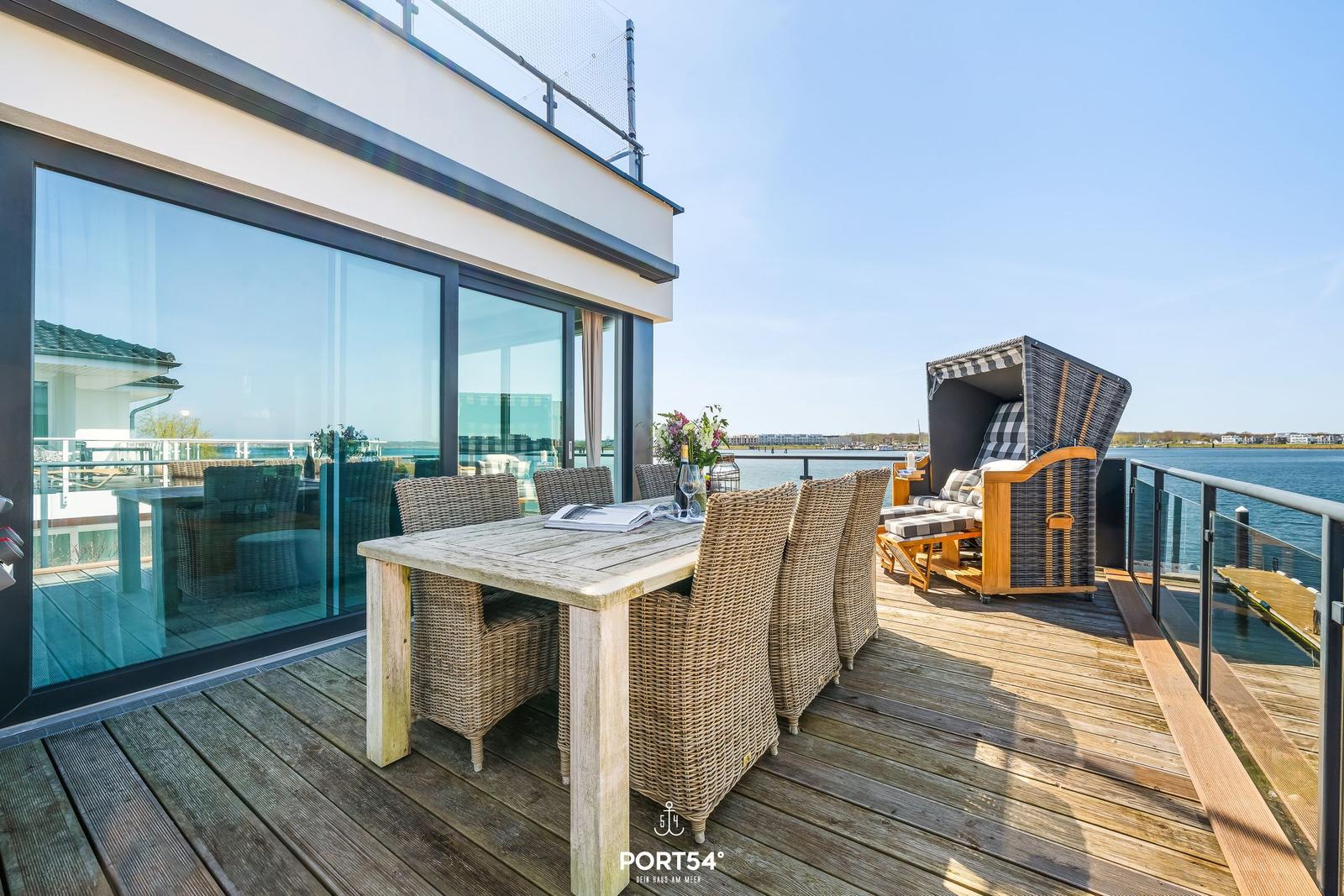 Terrasse mit Esstisch, Stühlen und Strandkorb am Wasser.