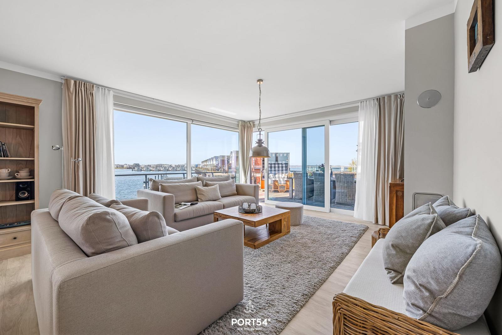 Wohnzimmer mit Blick auf Wasser und Terrasse. Möbel und Bodenbelag sind sichtbar.