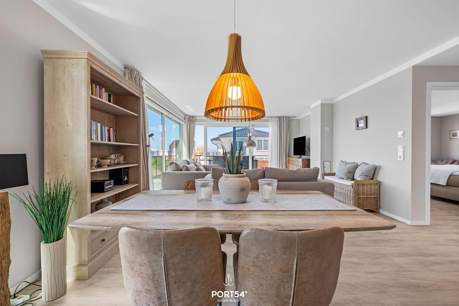 Essbereich mit Holztisch, Stühlen und einer großen Lampe. Im Hintergrund Wohnzimmer mit Sofa und Balkon.
