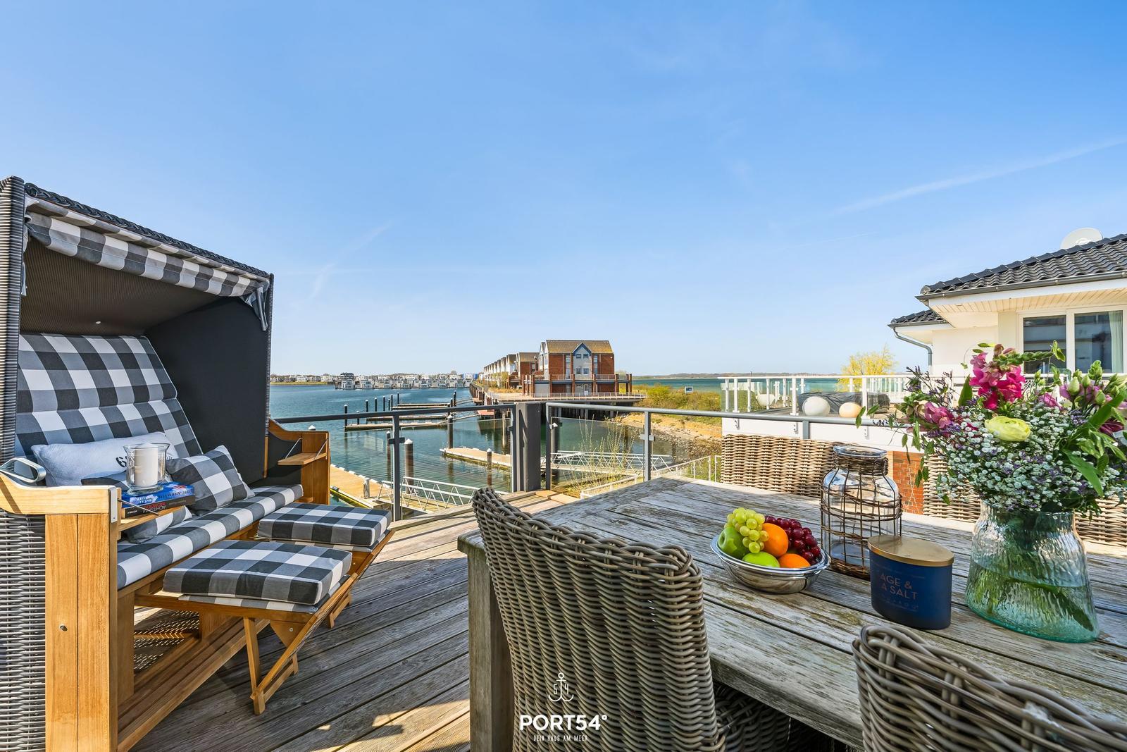 Terrasse mit Liege, Tisch und Früchten. Blick auf Wasser und Gebäude.