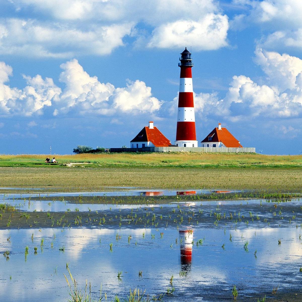 Red lighthouse with white stripes stands on an island. Water reflects it in the foreground.