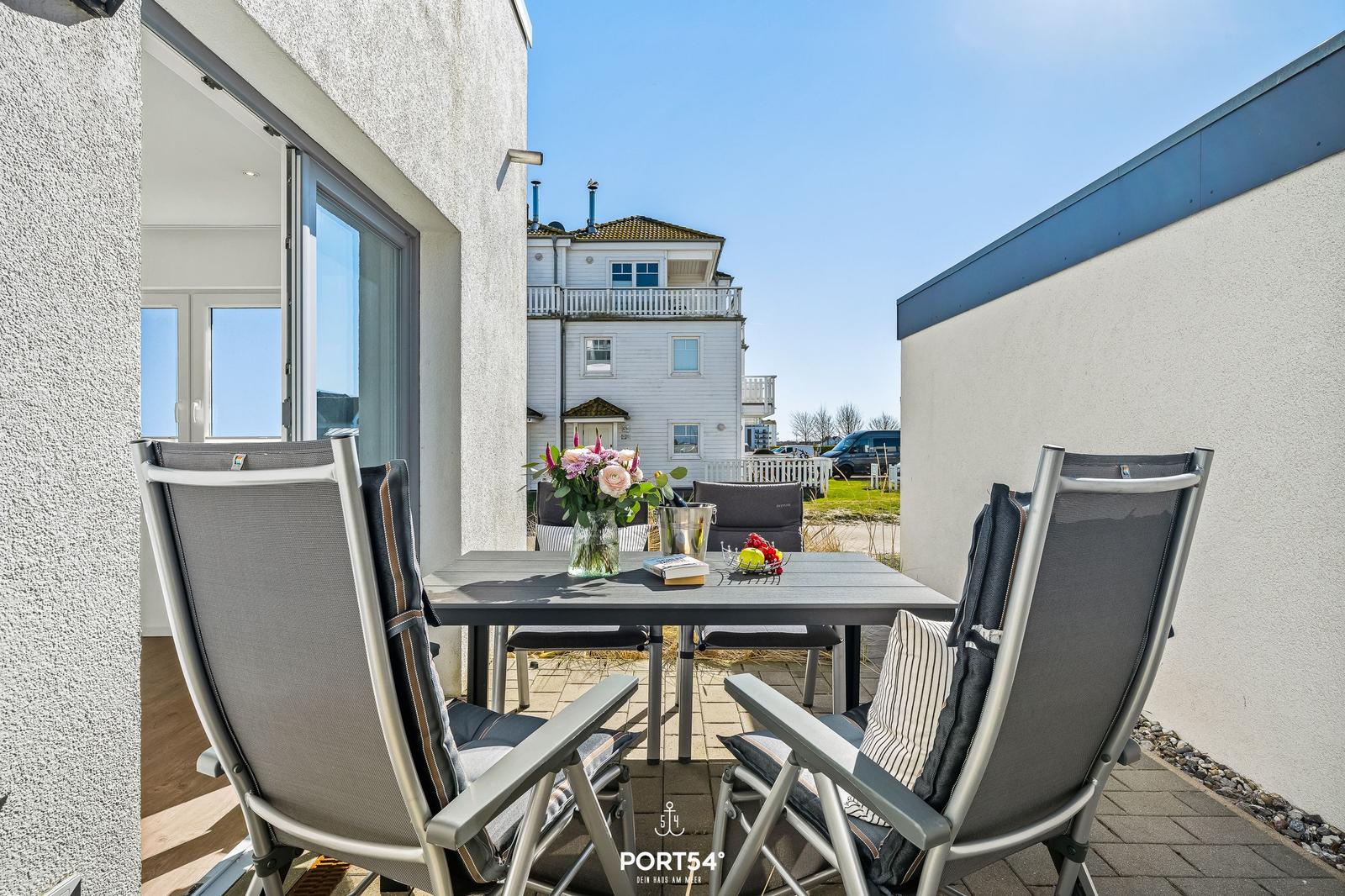 Terrasse mit Tisch und Stühlen, Blumen und Obst. Blick auf Haus und Himmel.