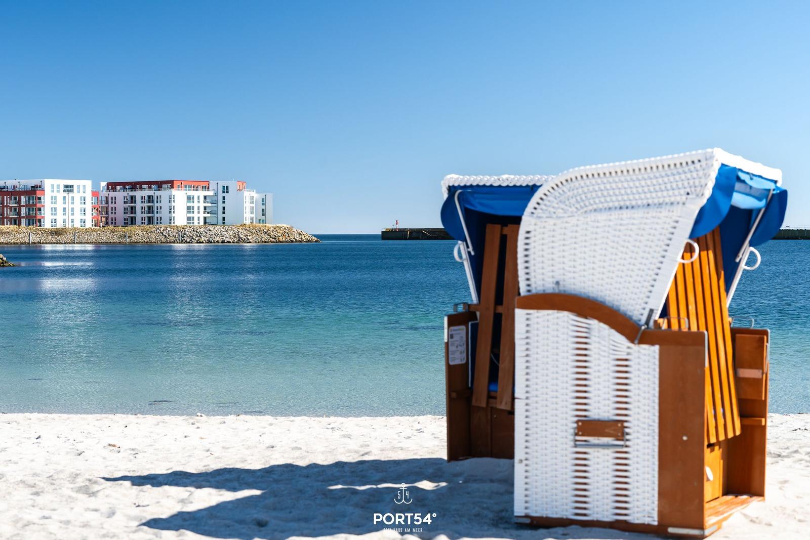 Strandkörbe stehen am sandigen Ufer, das Meer und moderne Gebäude sind im Hintergrund sichtbar.