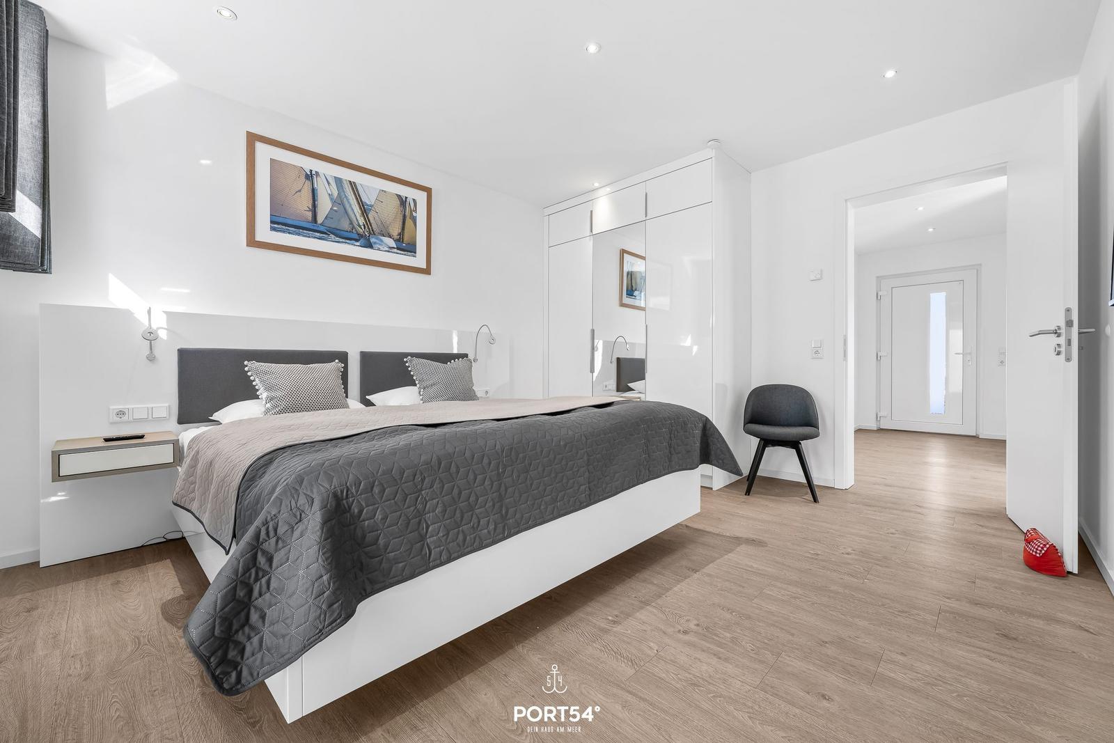 Schlafzimmer mit Doppelbett, Kleiderschrank und Stuhl. Holzboden und Wandbild sichtbar.