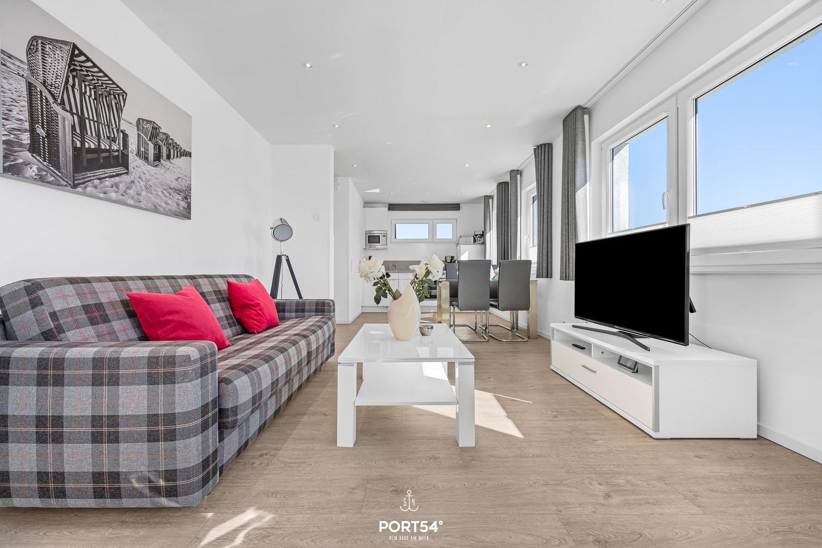 Wohnzimmer mit Sofa, Fernseher und großem Fenster. Essbereich im Hintergrund.
