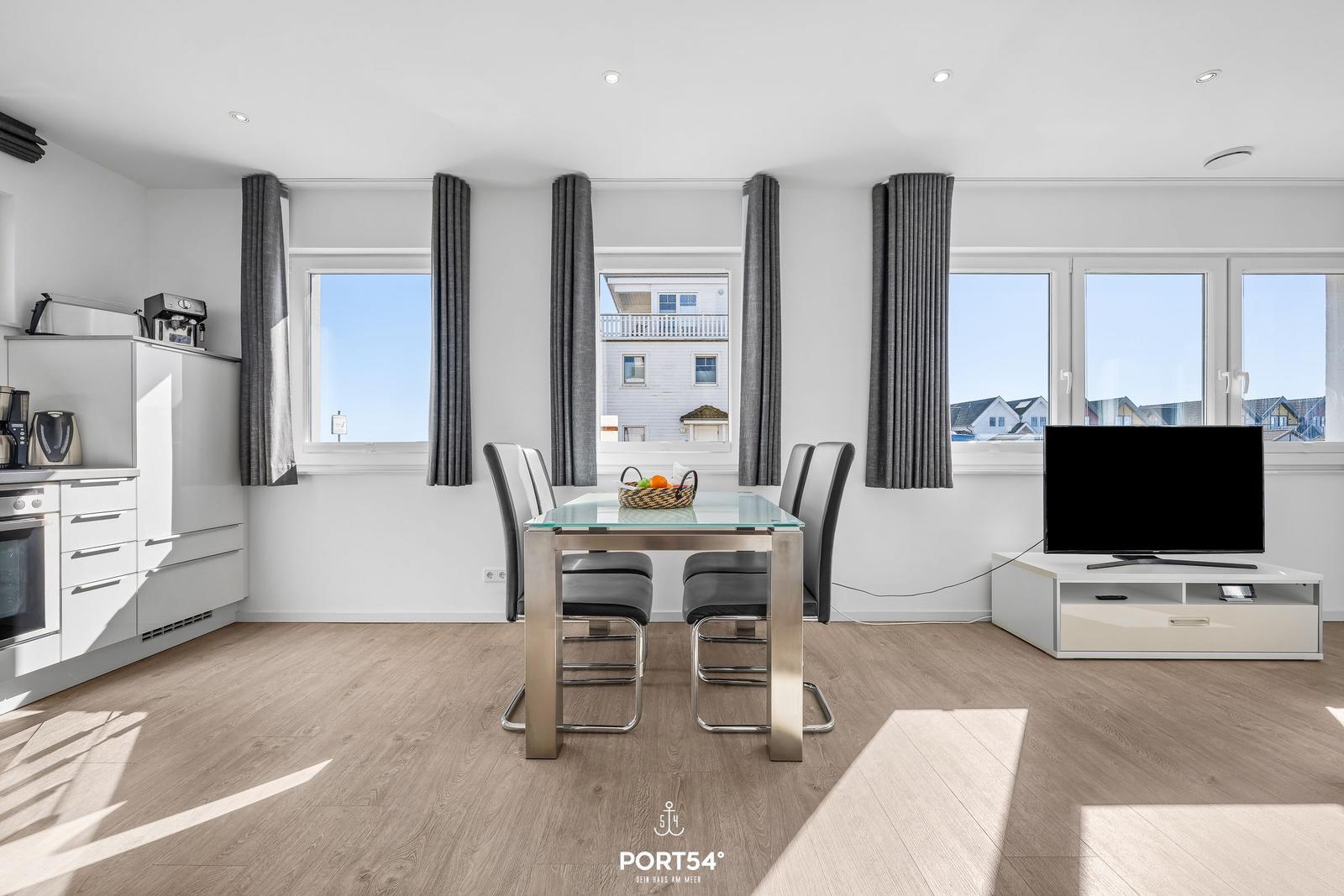 Essbereich mit Glasplatte, schwarzen Stühlen und TV. Fenster mit grauen Vorhängen.