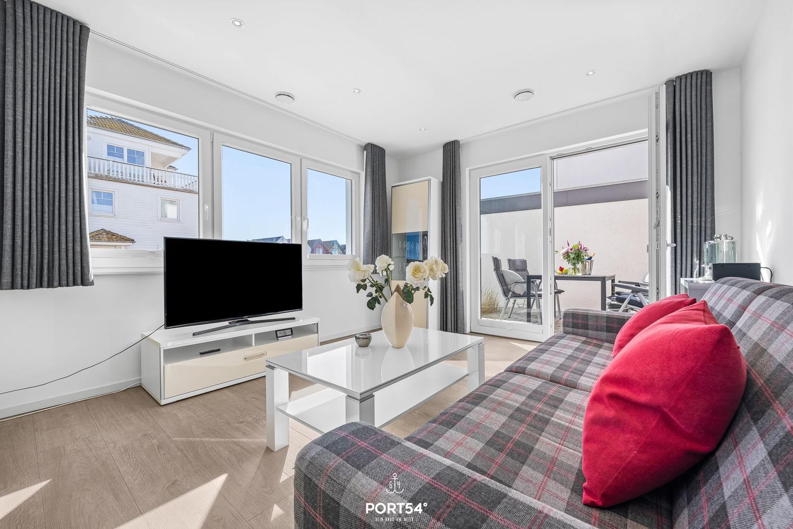 Wohnzimmer mit Sofa, Fernseher und Terrasse. Große Fenster und helle Wände.