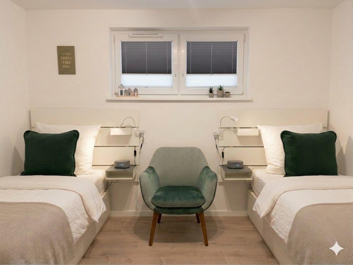 Two beds with green pillows, a chair, and a window with blinds.