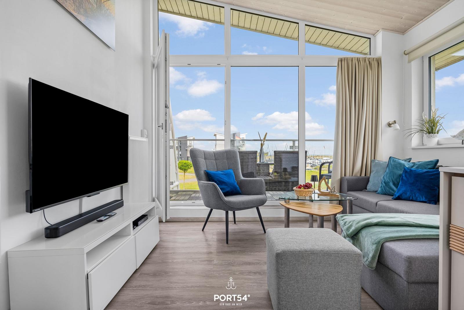 Wohnzimmer mit TV, Sofa und Blick auf Balkon. Große Fenster und helle Wände.