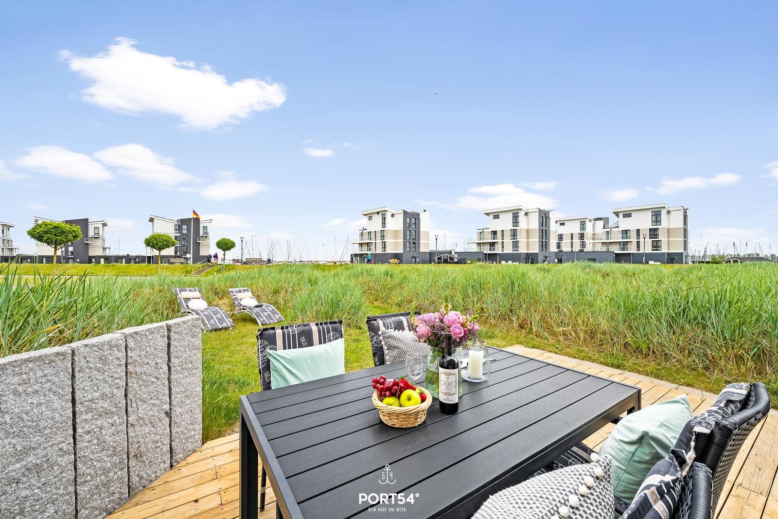 Terrasse mit Tisch, Stühlen und Obst. Im Hintergrund moderne Häuser und Grasland.