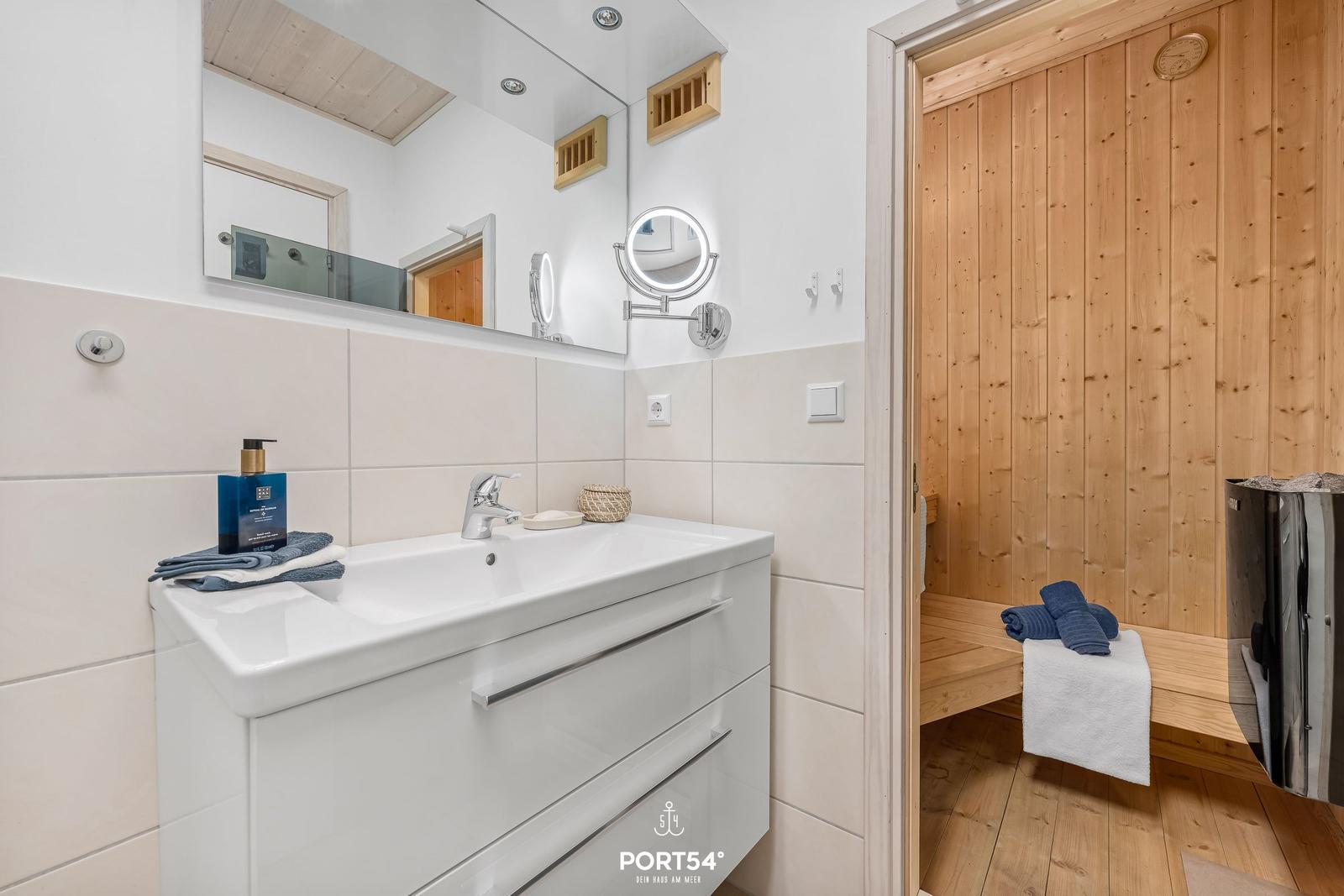 Bad mit Waschbecken, Spiegel und angrenzender Sauna mit Holzwand.